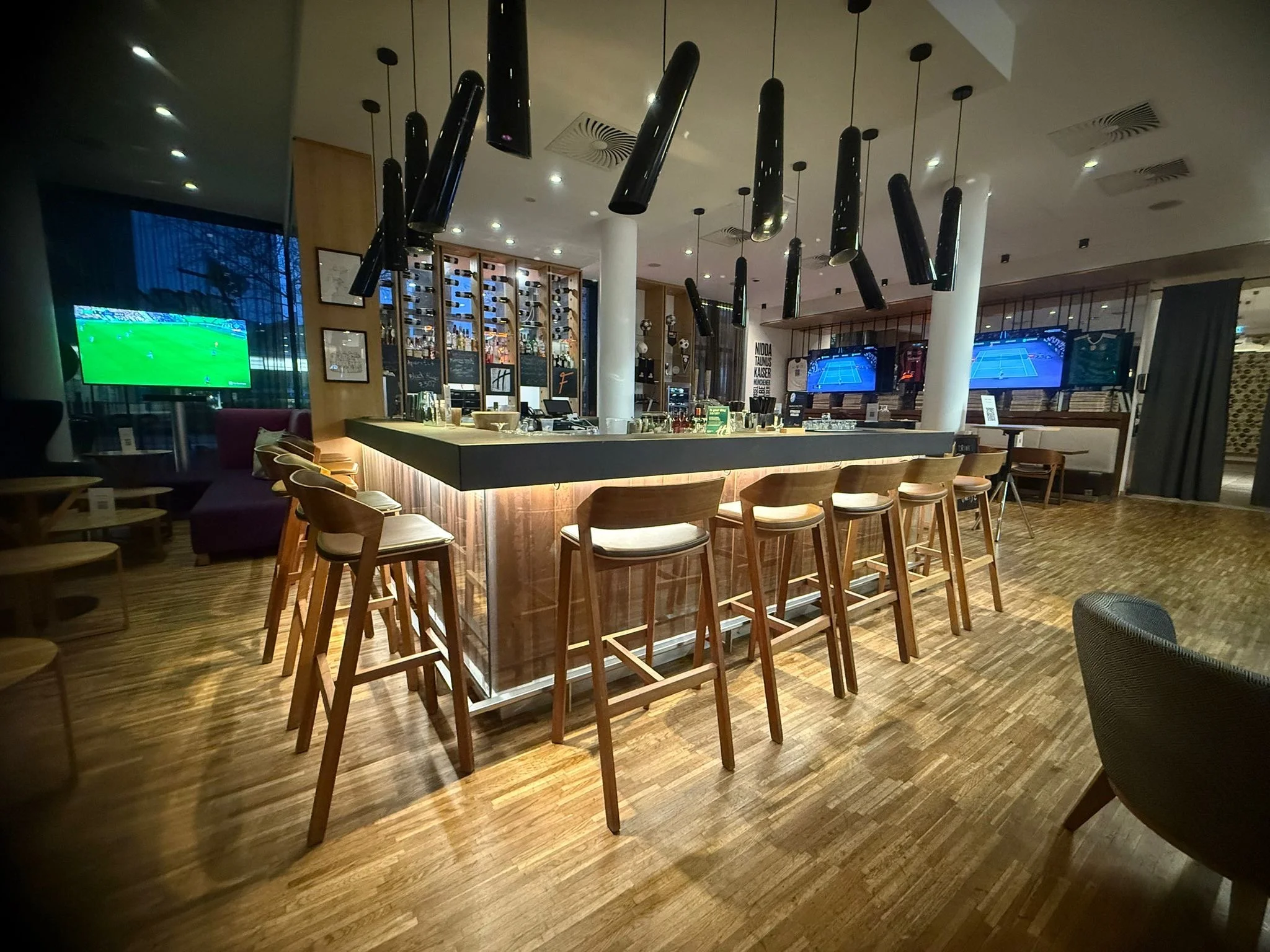 A modern bar interior with a high black bar counter, wooden bar stools with white cushions, and three ceiling-mounted black pendant lights. Multiple TVs display sports games, and a wall-mounted wine rack with bottles lines the back wall. The floor is