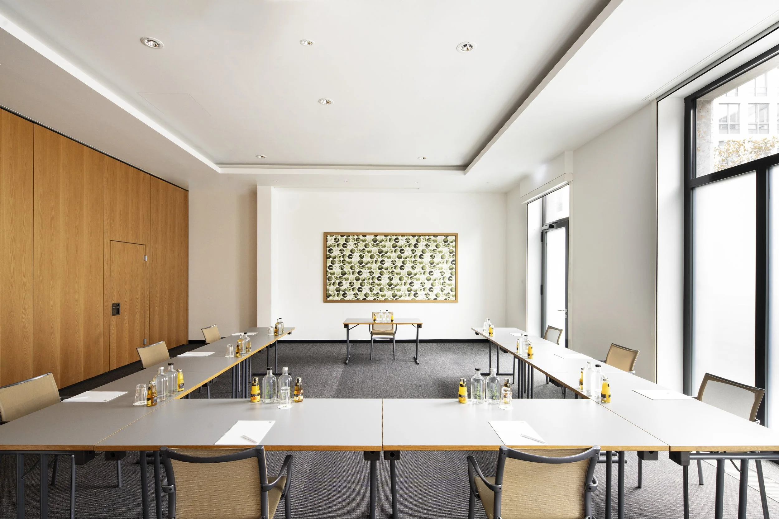 Empty conference room with tables arranged in U-shape, water bottles and notepads on tables, large windows, white walls, a framed artwork, and a wooden wall panel.