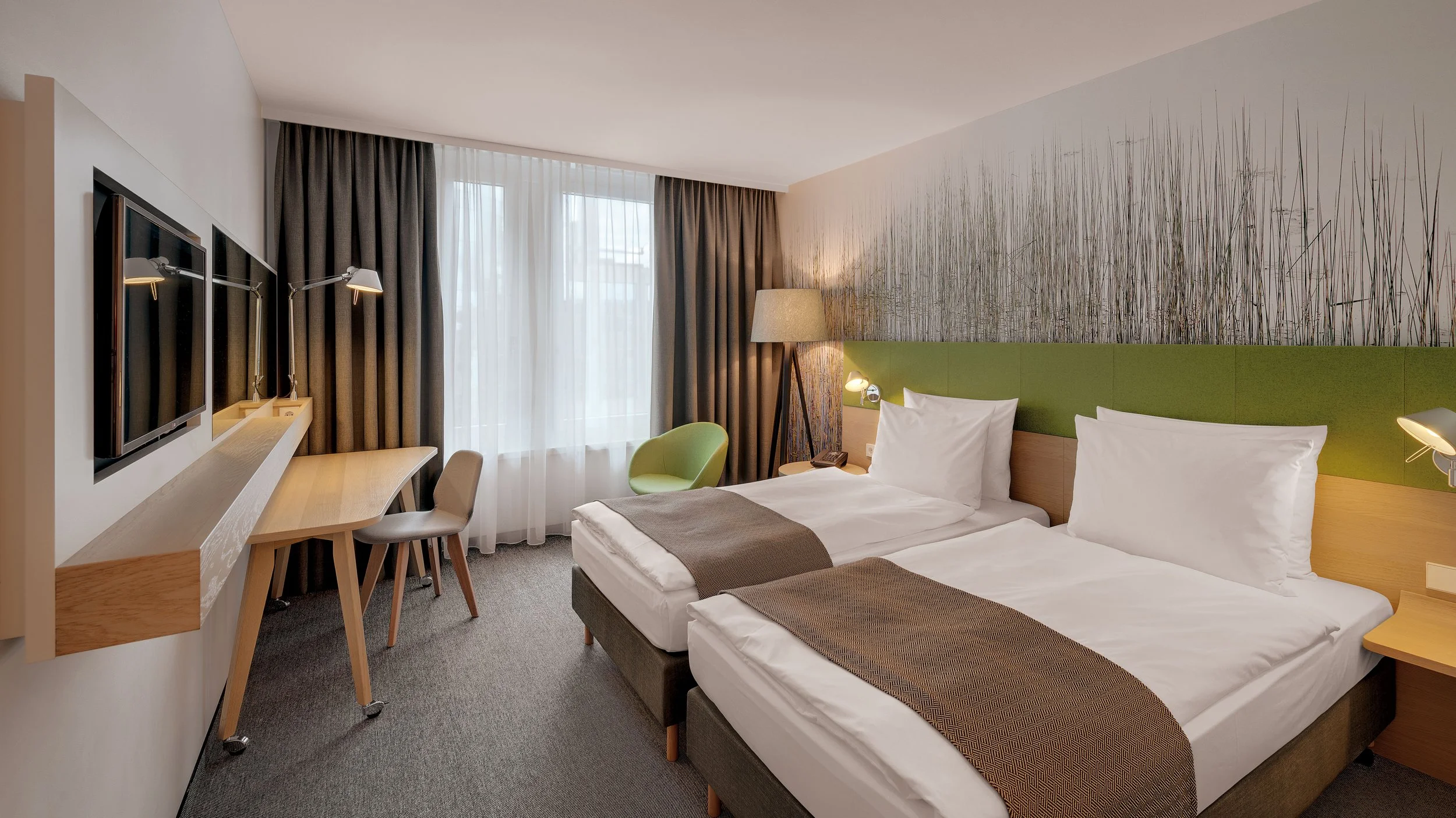 Modern hotel room with two twin beds, green headboard, bedside lamps, a flat-screen TV, a desk with chairs, a chair near the window, and a wall mural of grass.