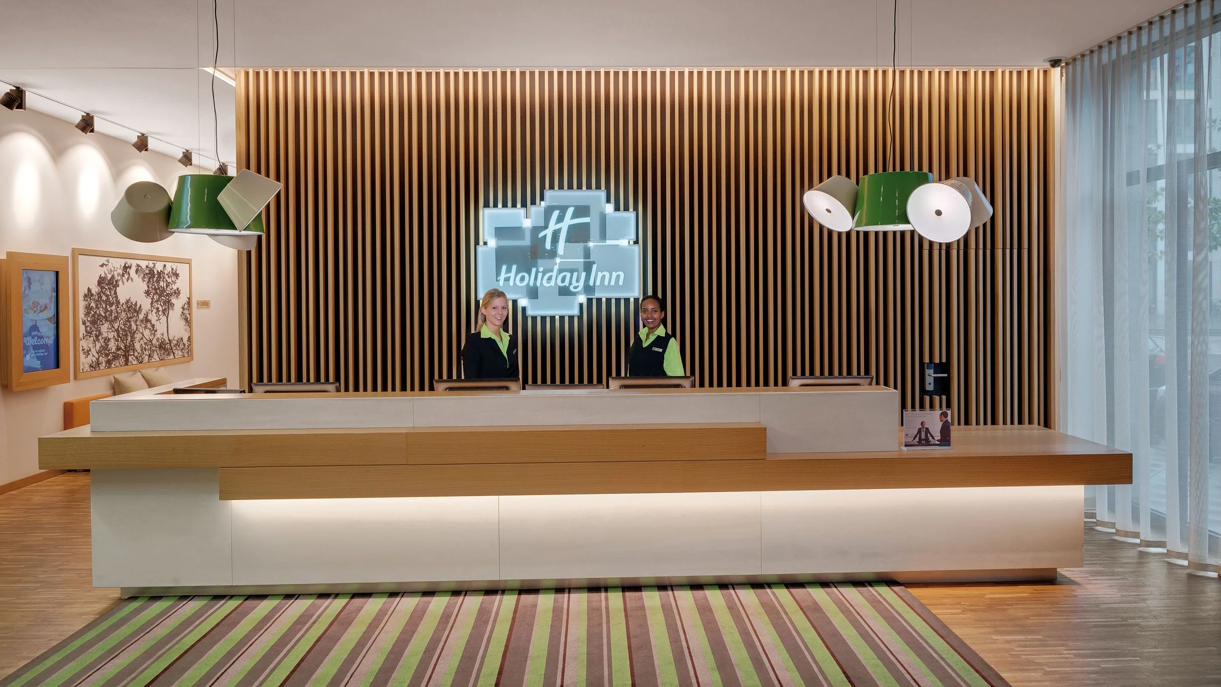 Front desk of a Holiday Inn hotel with two smiling staff members standing behind the counter, a neon Holiday Inn sign on a wooden slat accent wall, modern pendant lights, and a window with curtains.