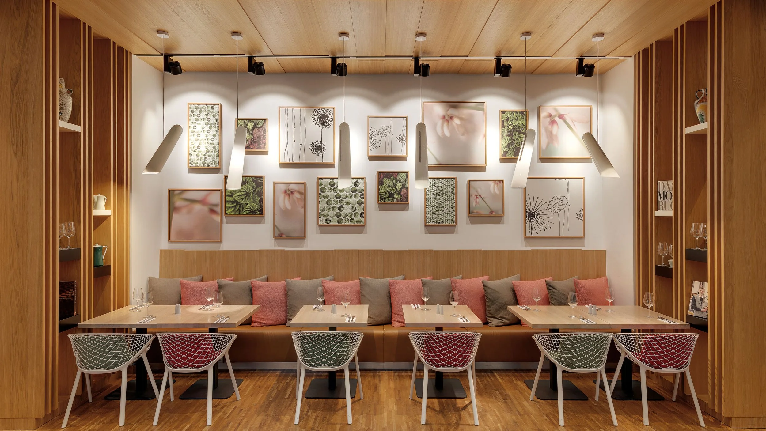 A cozy dining area in a modern restaurant featuring a wall with framed floral artwork, pendant lighting, wooden shelving on both sides holding decorative items, a row of cushions on a bench, and tables set with wine glasses and cutlery.