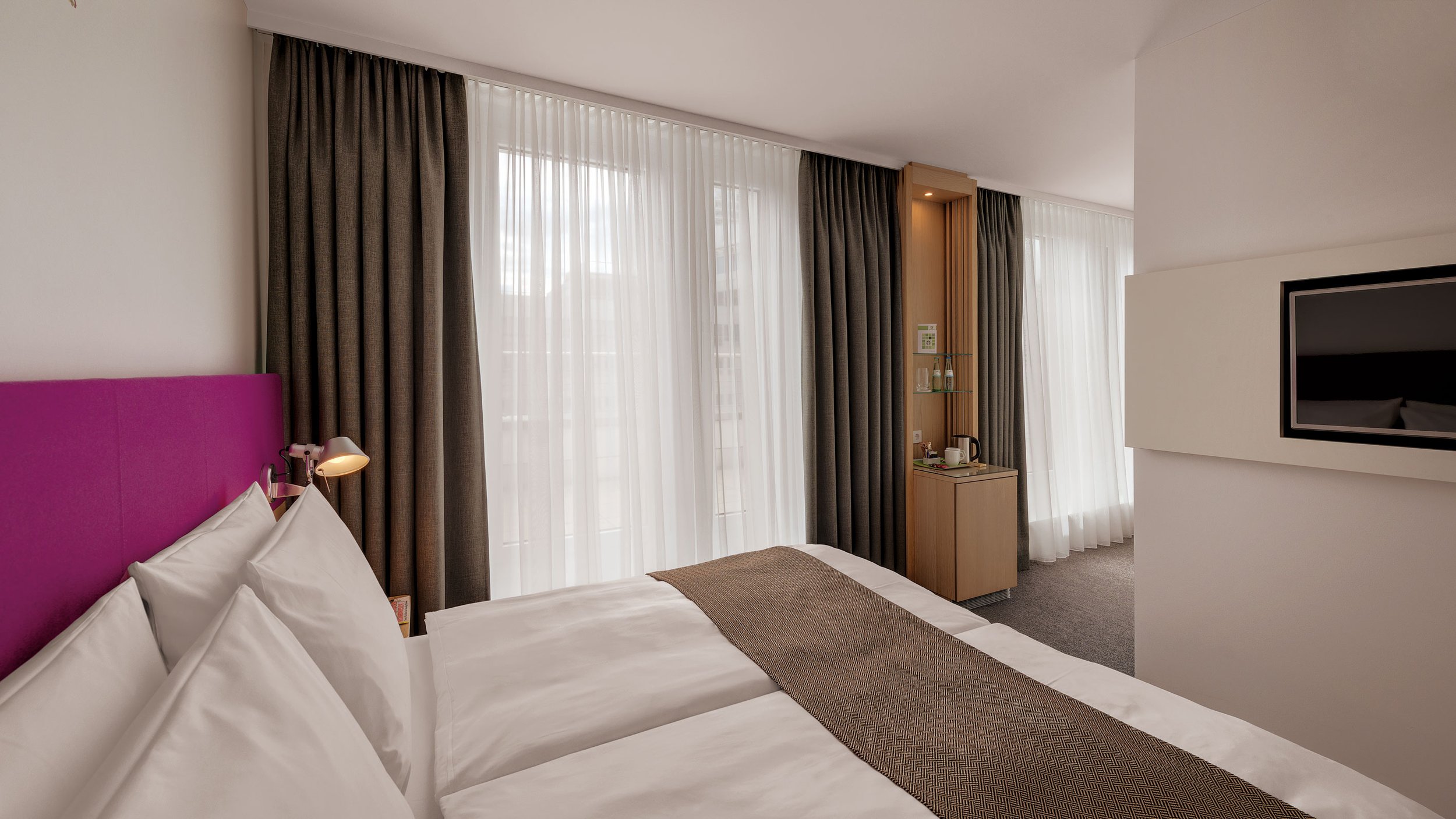 Hotel room with bed, pink headboard, white pillows, bedside lamp, large window with sheer white curtains and gray blackout curtains, wooden cabinet with tea and coffee making facilities, visible television, light-colored walls, neutral-colored carpet.