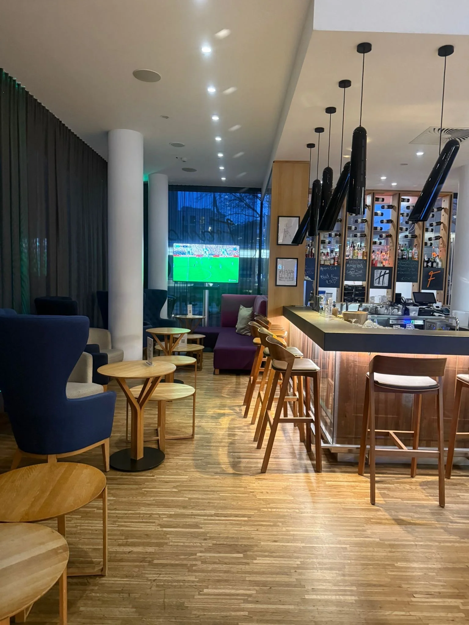 A cozy bar and lounge area with a wooden floor, a bar counter with black pendant lights, seating with armchairs and bar stools, a large window with curtains, and a television screen showing a sports game.