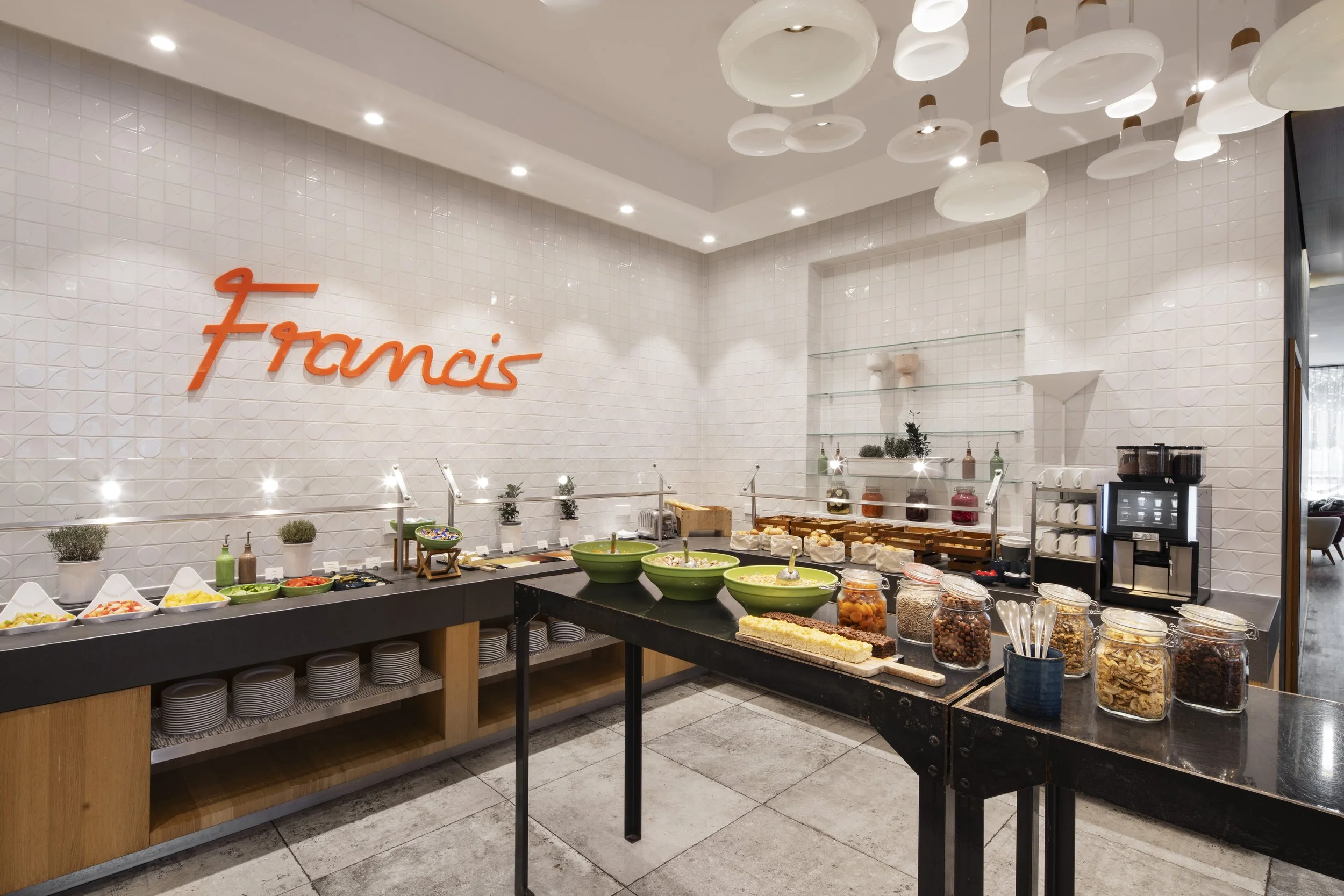 Buffet table with various salad toppings, jars of dried fruit and nuts, and a cake, in a modern restaurant interior with white tile walls and decorative lighting.