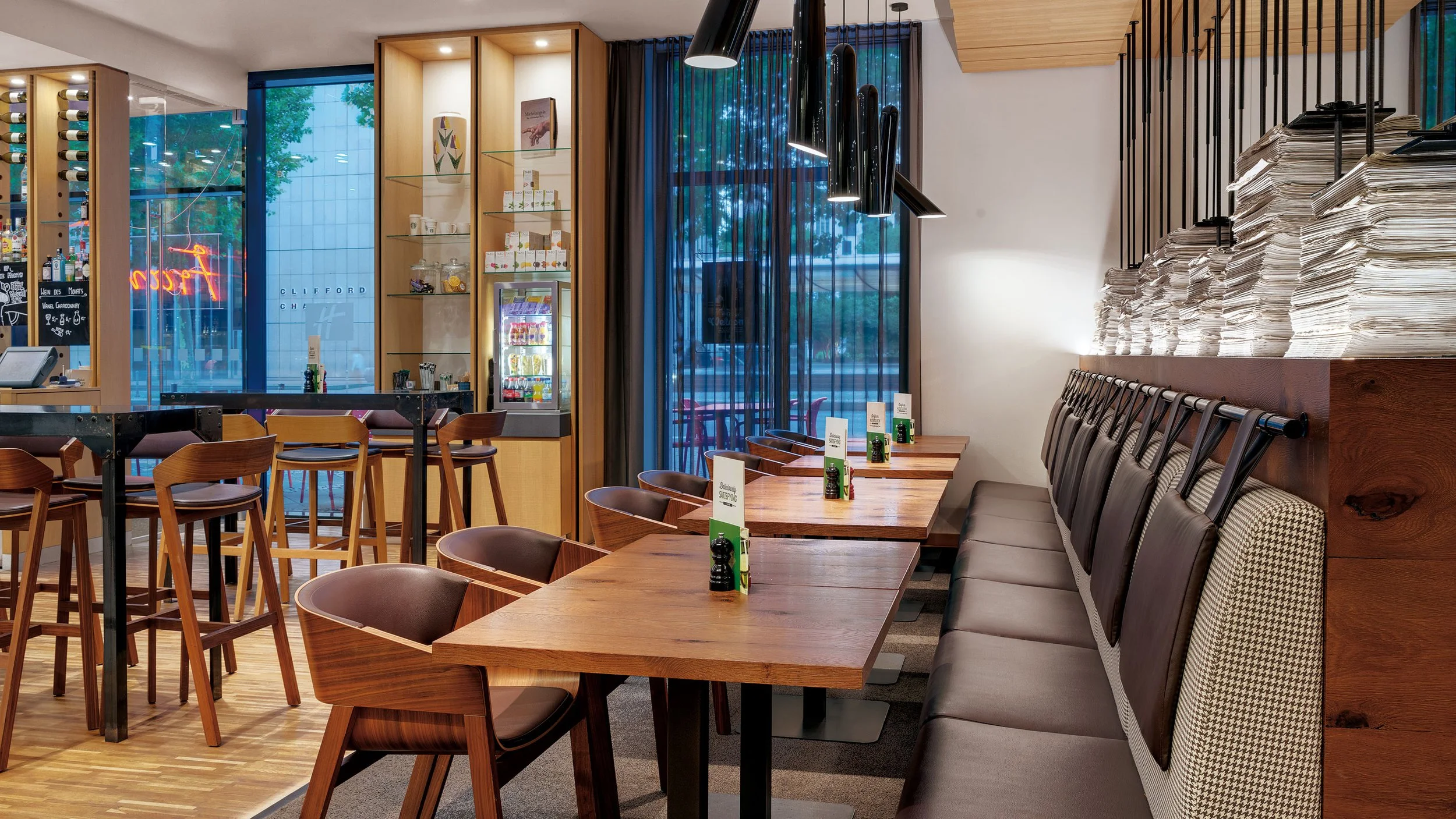 Interior of a modern restaurant with wooden tables and chairs, a long cushioned bench, large windows with black curtains, and stacks of newspapers on a wall.