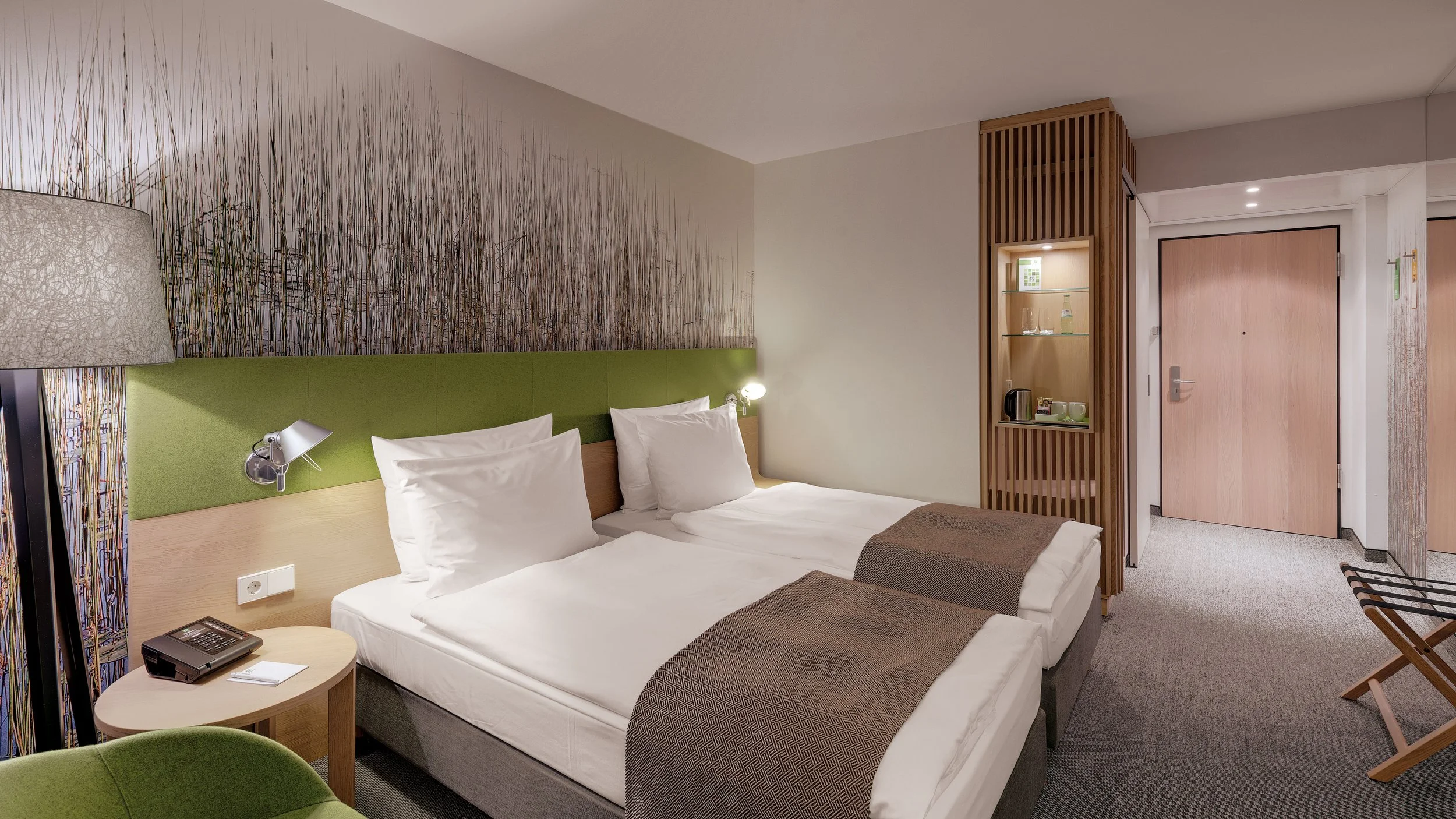 A hotel room with two beds, a nightstand with a phone, a bedside lamp, and a decorative wall panel of tall grass design. There's a wooden partition with glass shelves holding glasses and a water bottle, and an entry door in the background.