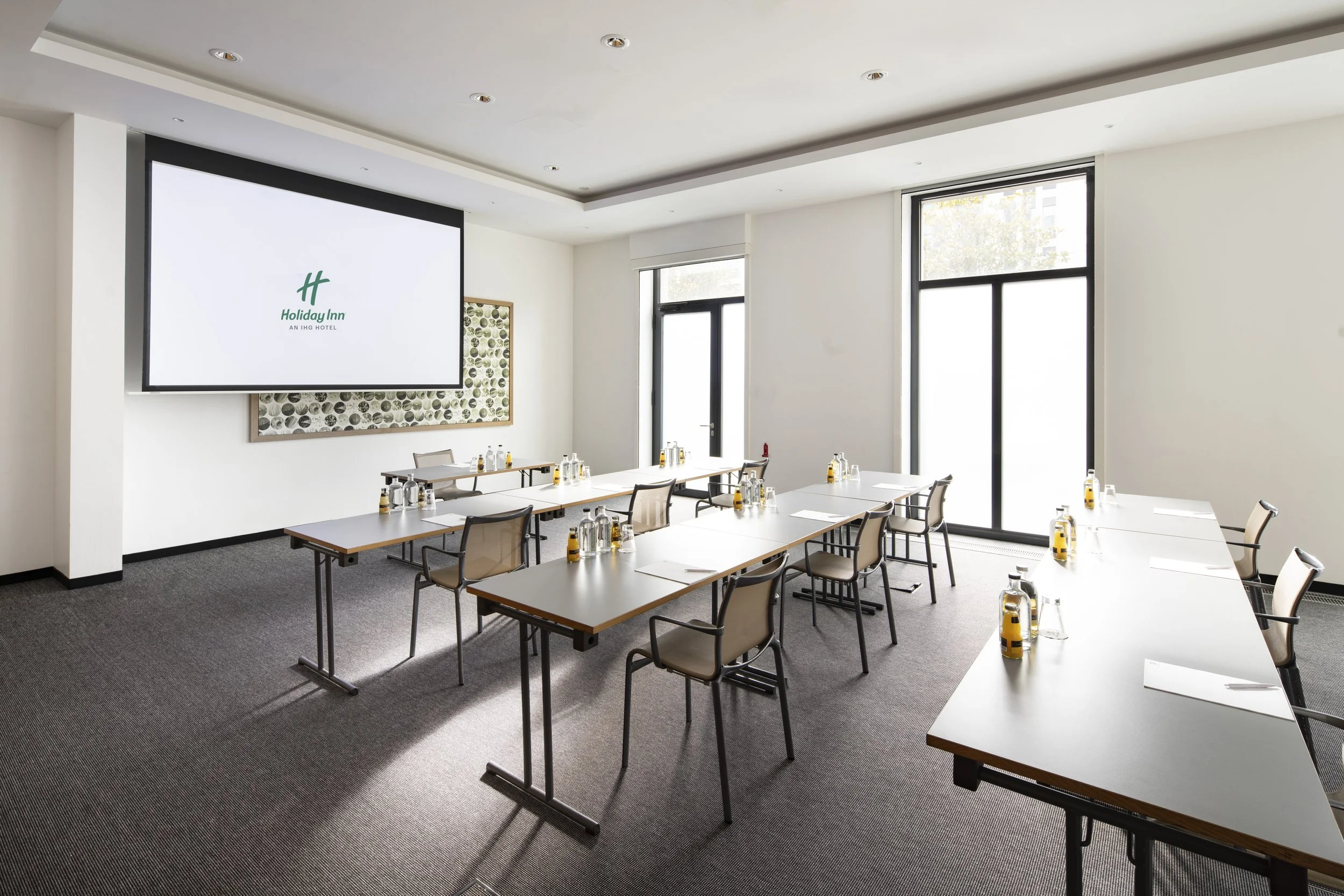 A conference room set up for a meeting with white tables, beige chairs, water bottles, glasses, and notepads. There is a large screen on the wall displaying the Holiday Inn logo. The room has large windows letting in natural light.