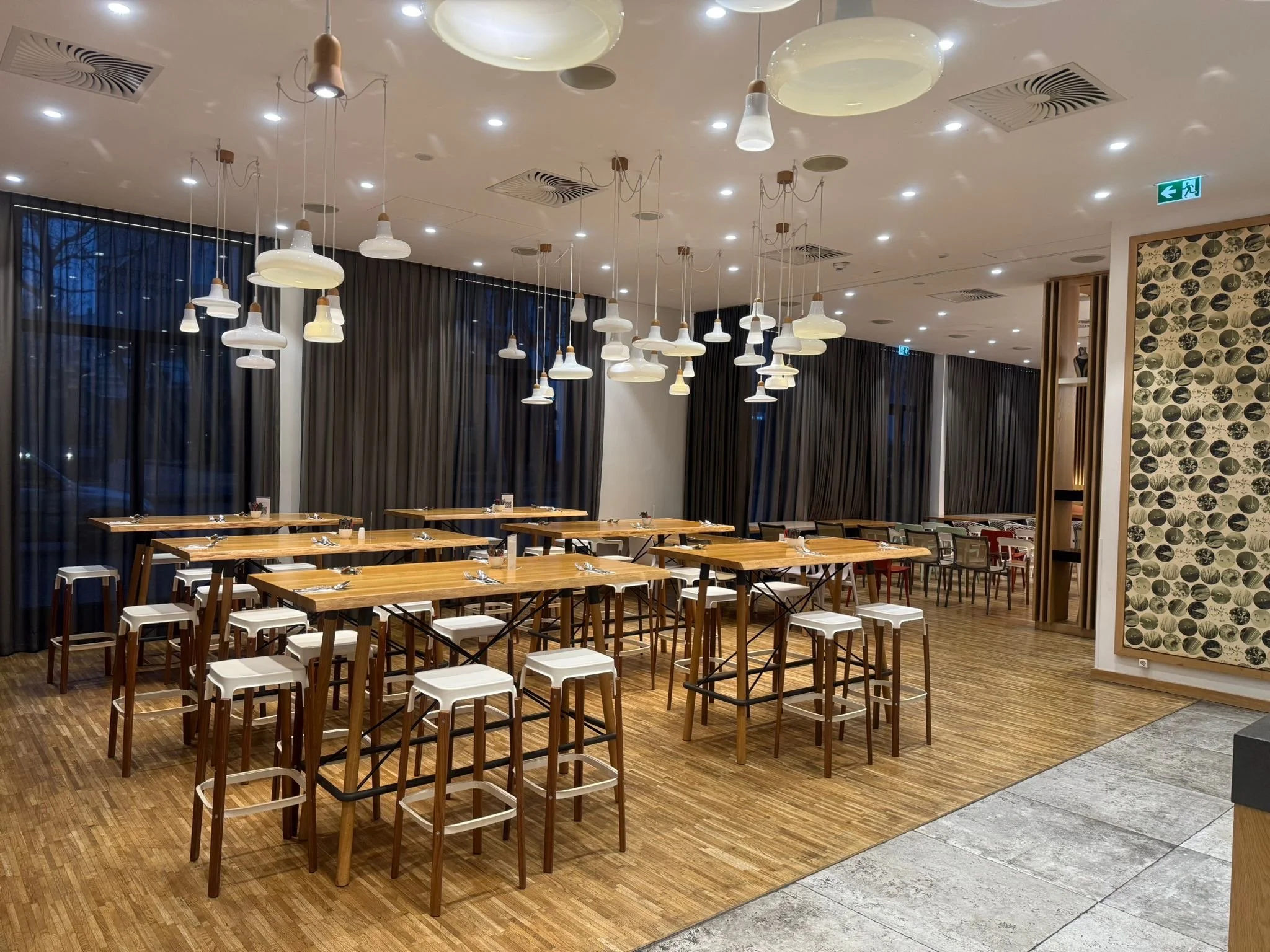 Interior of a modern restaurant or cafe with wooden tables and white stools, large windows with black curtains, numerous hanging white pendant lights, and a decorative wall with circular patterns.