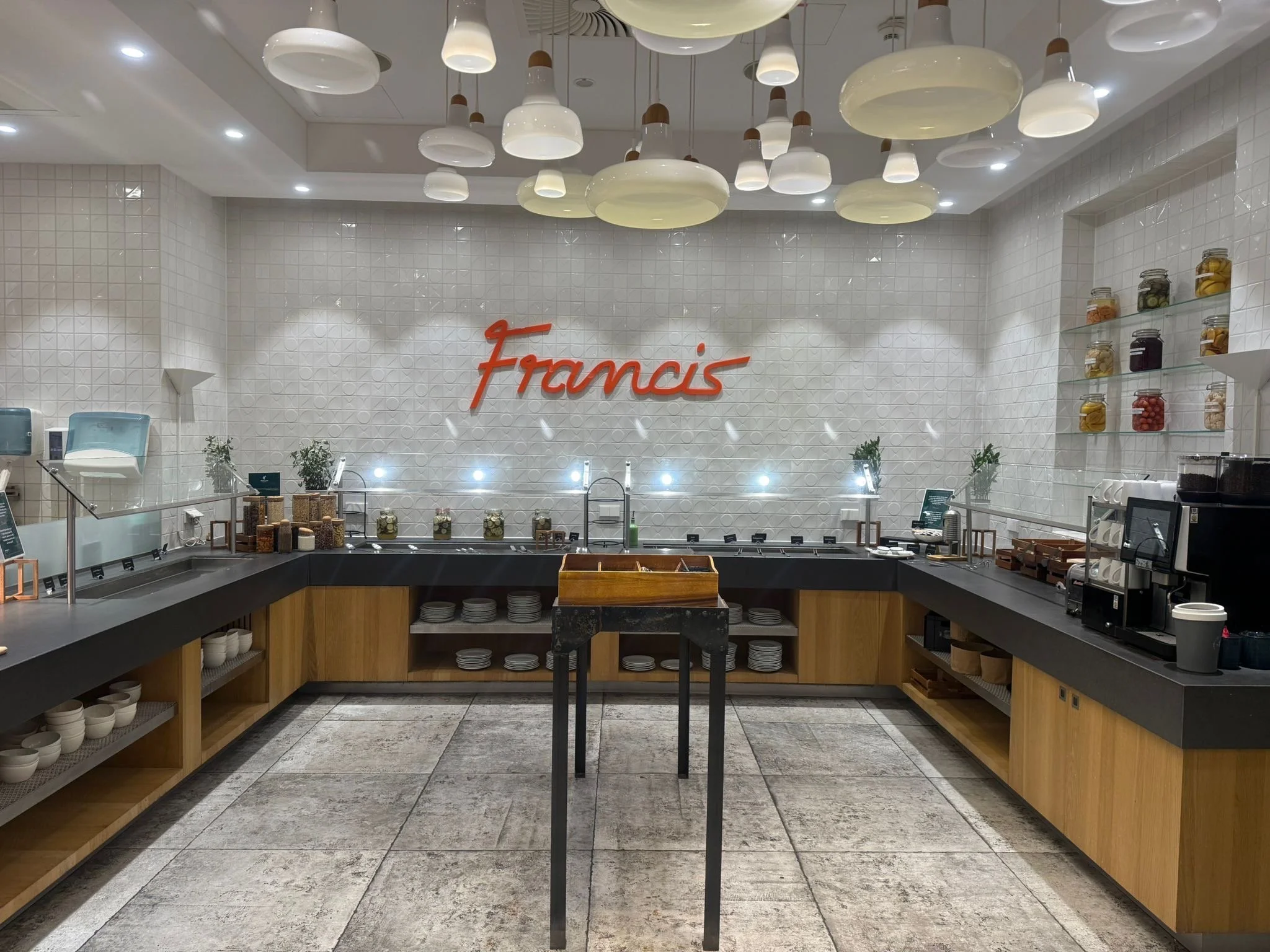 A cafeteria or food court counter with a white textured wall featuring red cursive 'Francis' sign, white pendant lights on the ceiling, glass jars with food items on the right, and shelves with bowls and plates below. The counter has a black surface 