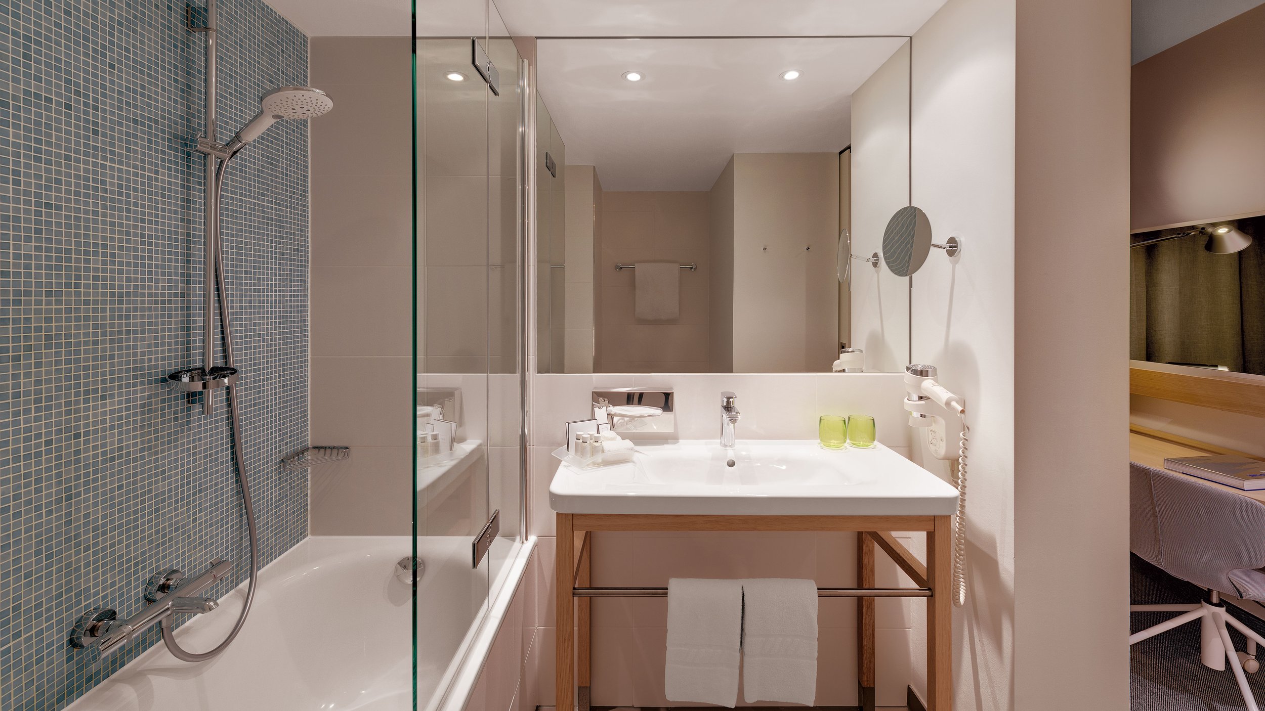 Modern hotel bathroom with walk-in shower with mosaic tiles, white bathtub, mirror, vanity with sink, two green glasses, wall-mounted hairdryer, towel rack with towel, and a small desk with chair in living area.