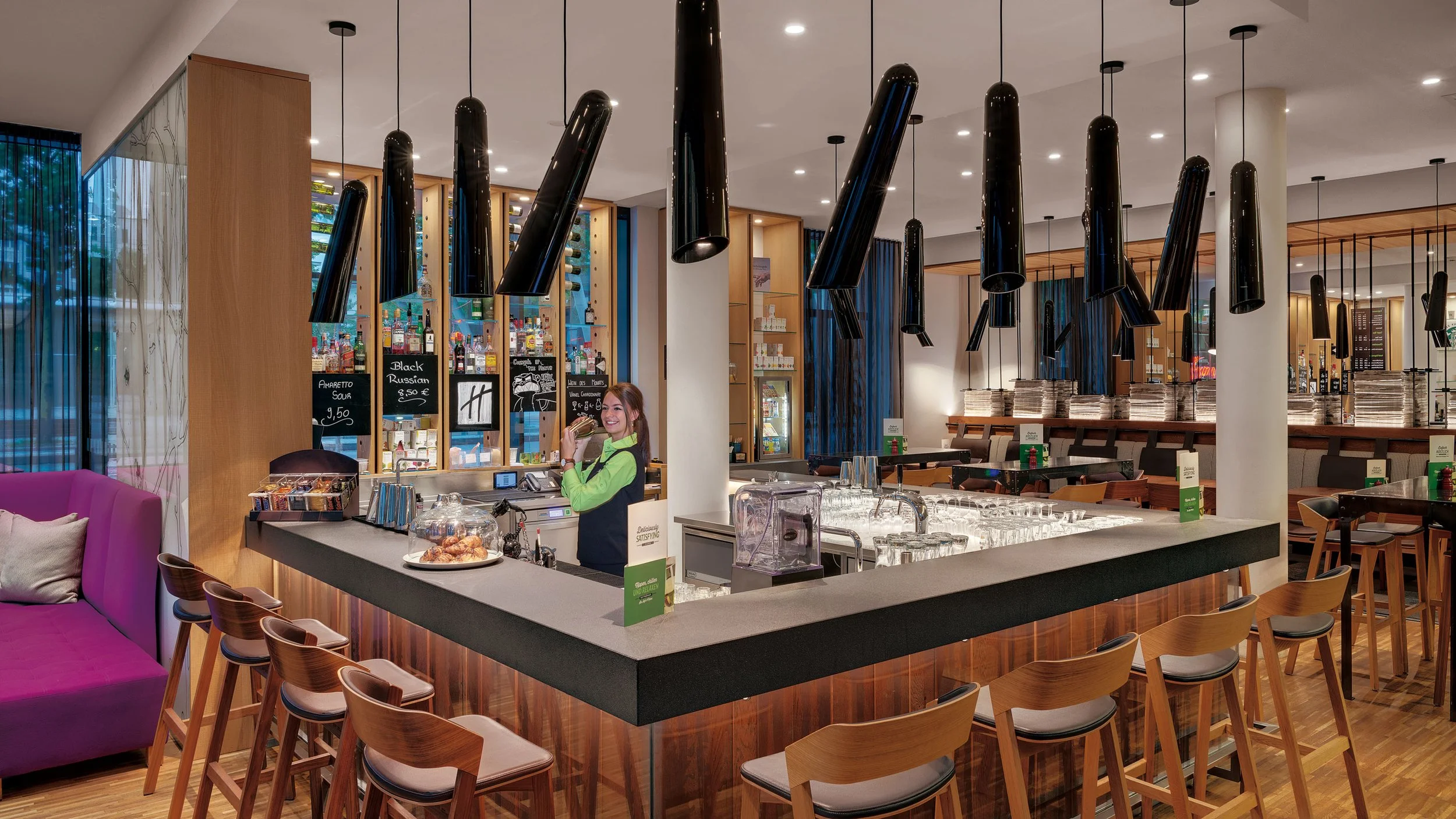 Interior of a modern bar or restaurant with a woman behind the counter, black pendant lights hanging from the ceiling, wine bottles displayed on shelves, a purple sofa with cushions, and a dining area with tables and chairs.