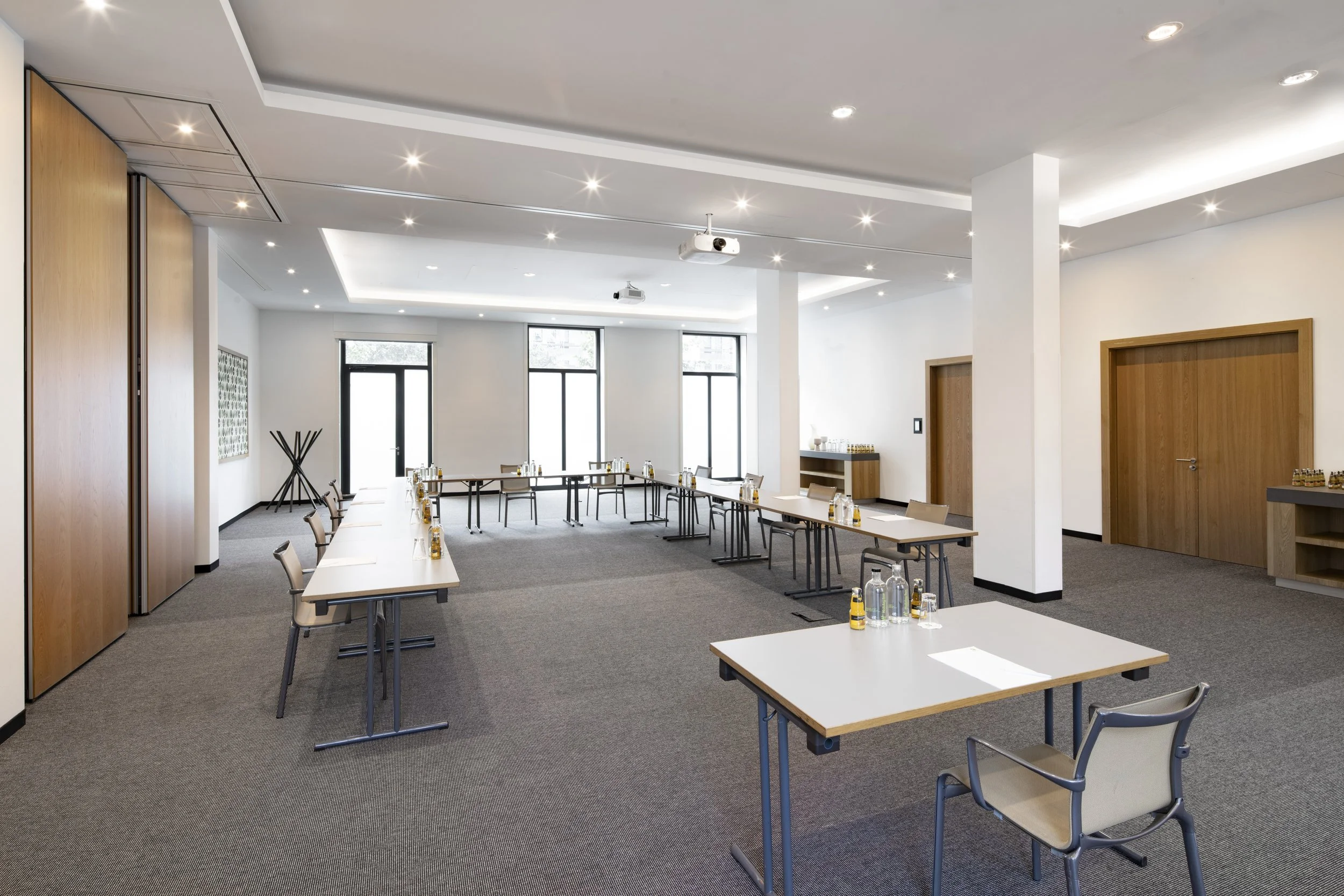 A conference room with white walls, large windows, and ceiling lights. There are long tables arranged in a U-shape with water bottles and glasses. Wooden doors and a small shelf with items are visible.