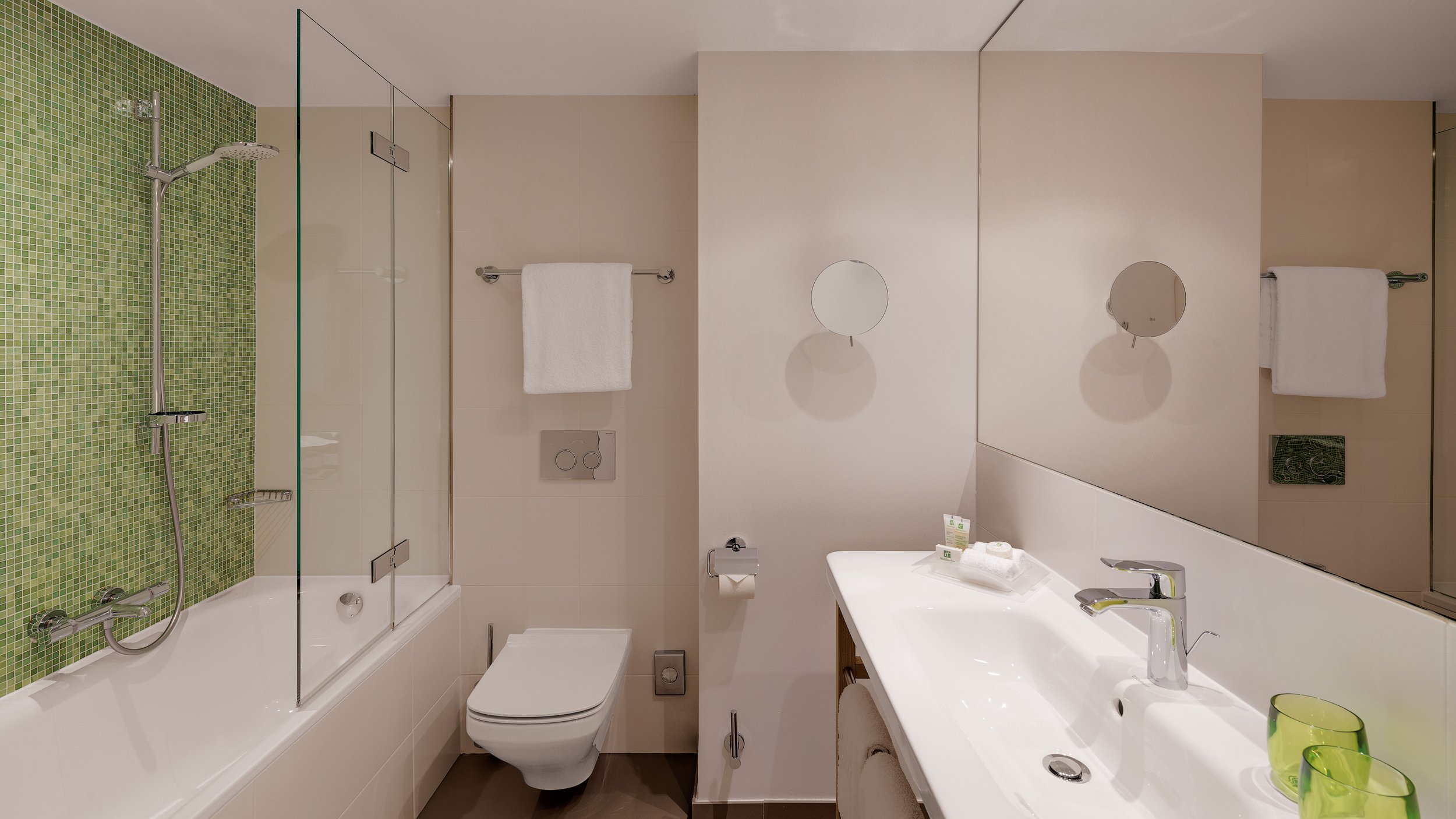 Modern bathroom with a shower area featuring green mosaic tiles, a glass shower door, a white sink with a faucet, a mirror, a toilet with a dual-flush button, and towels hanging on a rail. The decor is minimalistic and clean.