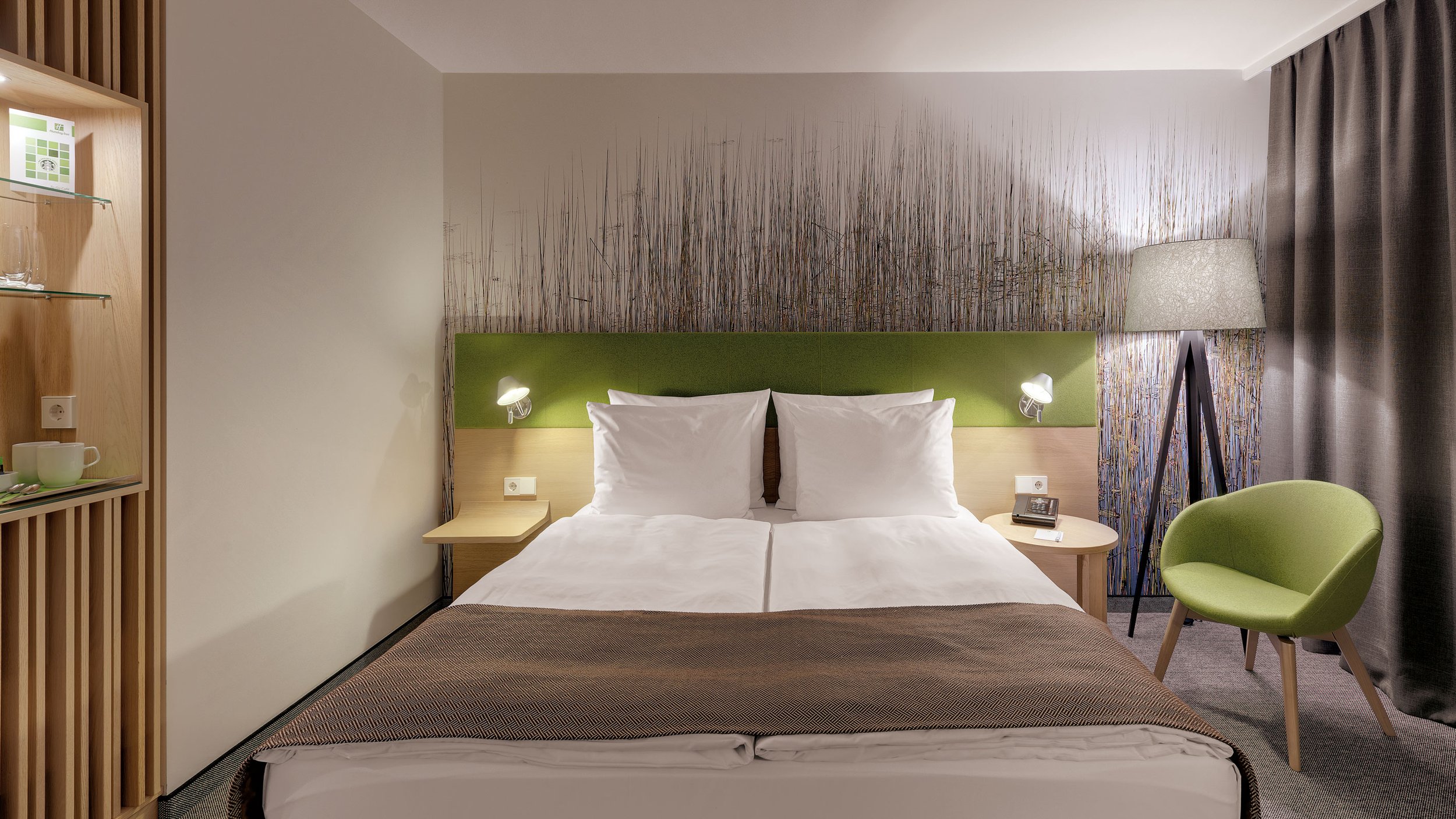 A hotel room with a bed, nightstand, and modern decor. The bed has white linens, pillows, and a green headboard. There are wall-mounted reading lights, a lamp on a standing tripod, and a green chair near a window with curtain.