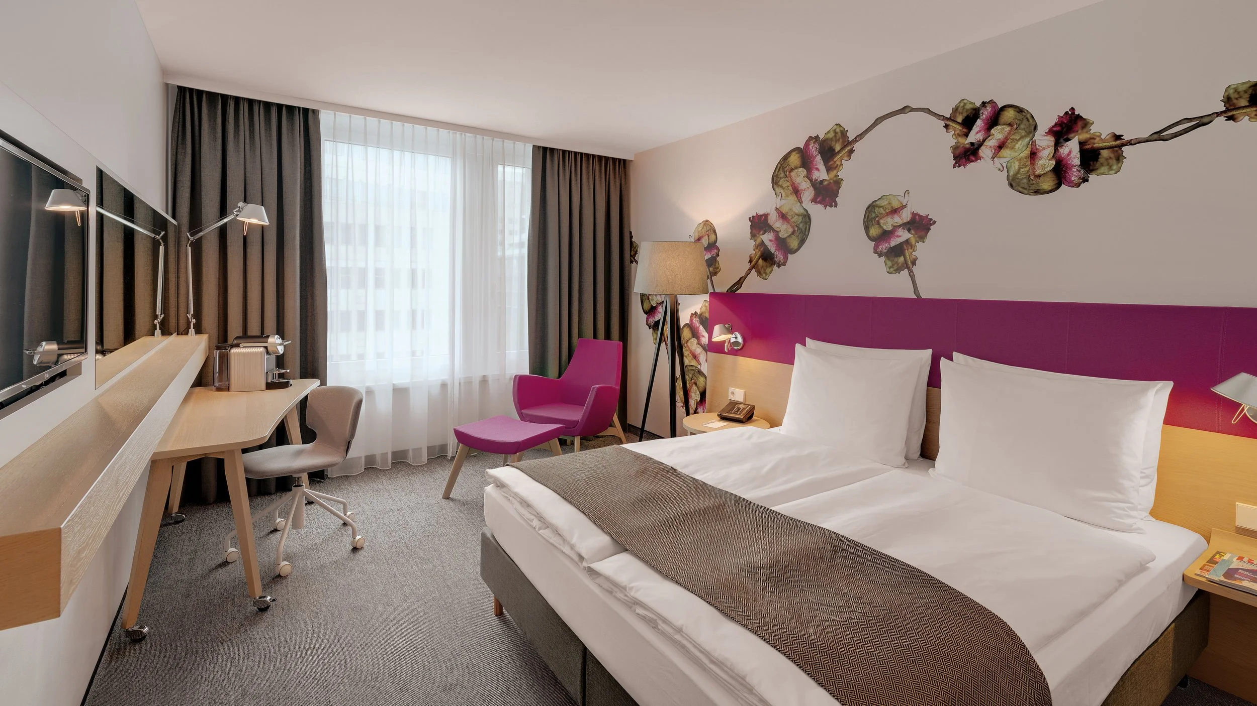 A hotel room with a bed that has white sheets and pillows, a pink headboard, a colorful floral mural on the wall behind the bed, a purple armchair with a matching ottoman, a wooden desk with a beige chair, a tall floor lamp, curtains covering a large