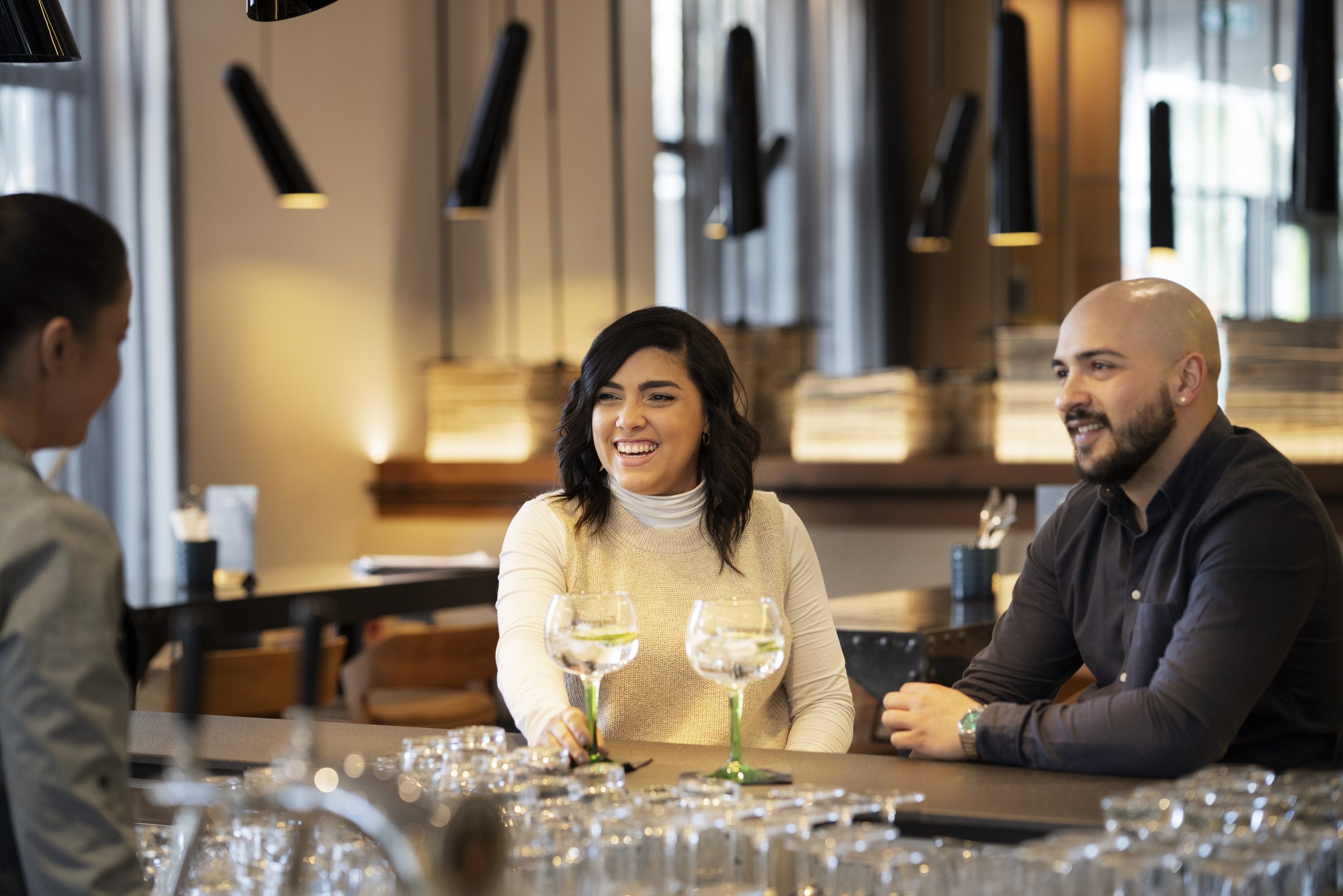 Three people sitting at a restaurant table, smiling and chatting, with glasses of water and wine glasses in front of them.