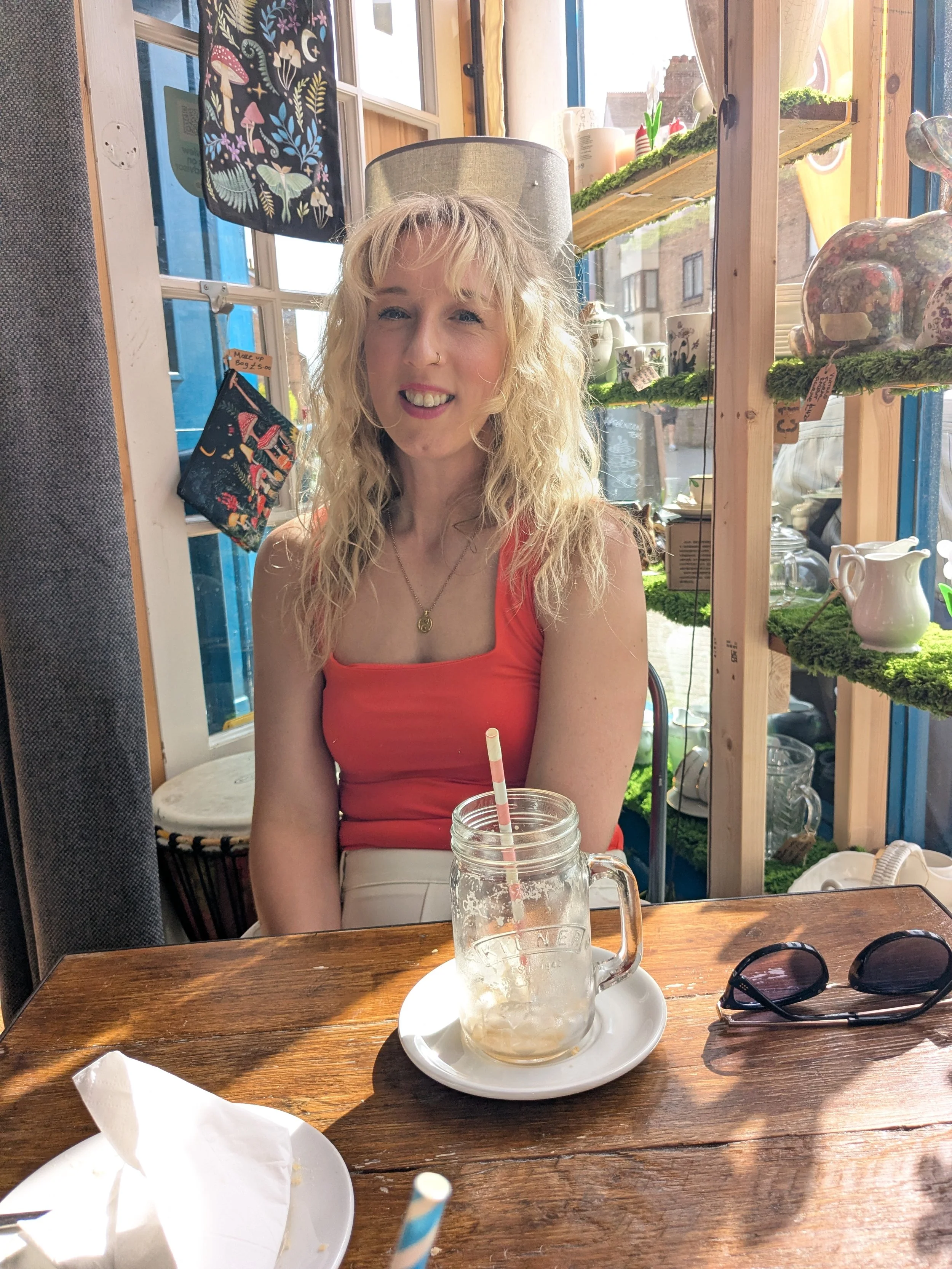 A woman with blonde curly hair, wearing an orange tank top, sitting at a wooden table inside a café, smiling at the camera. There is a glass jar with a straw and a partially eaten dessert on a plate in front of her. Sunglasses are on the table, and the café has decorative shelves with ceramics and plants.