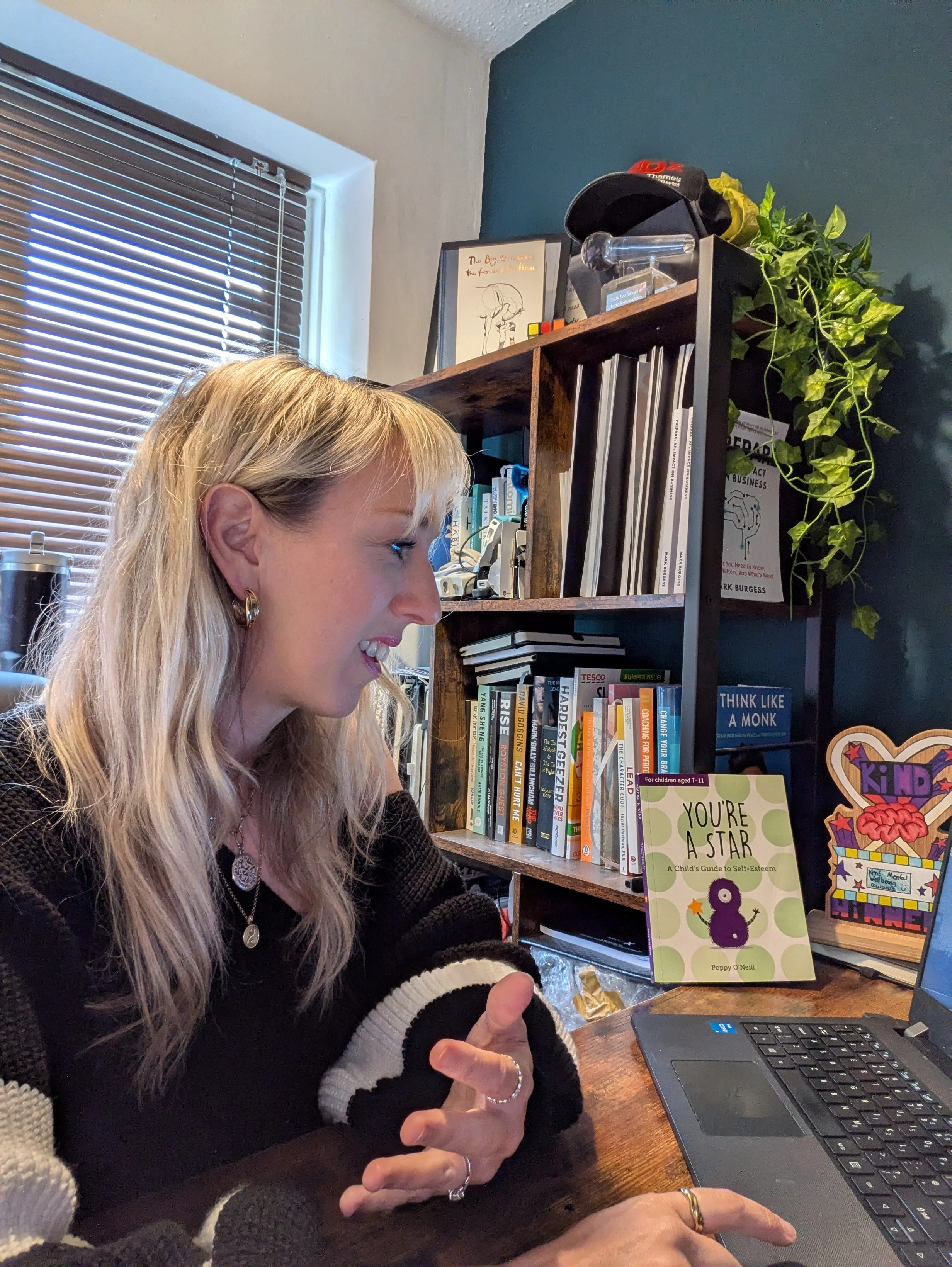 A woman with long blonde hair wearing a black and white sweater, sitting at a desk, smiling and looking at a laptop. Behind her is a bookshelf with books, a framed drawing, and a large plant, with a window with blinds on the left.