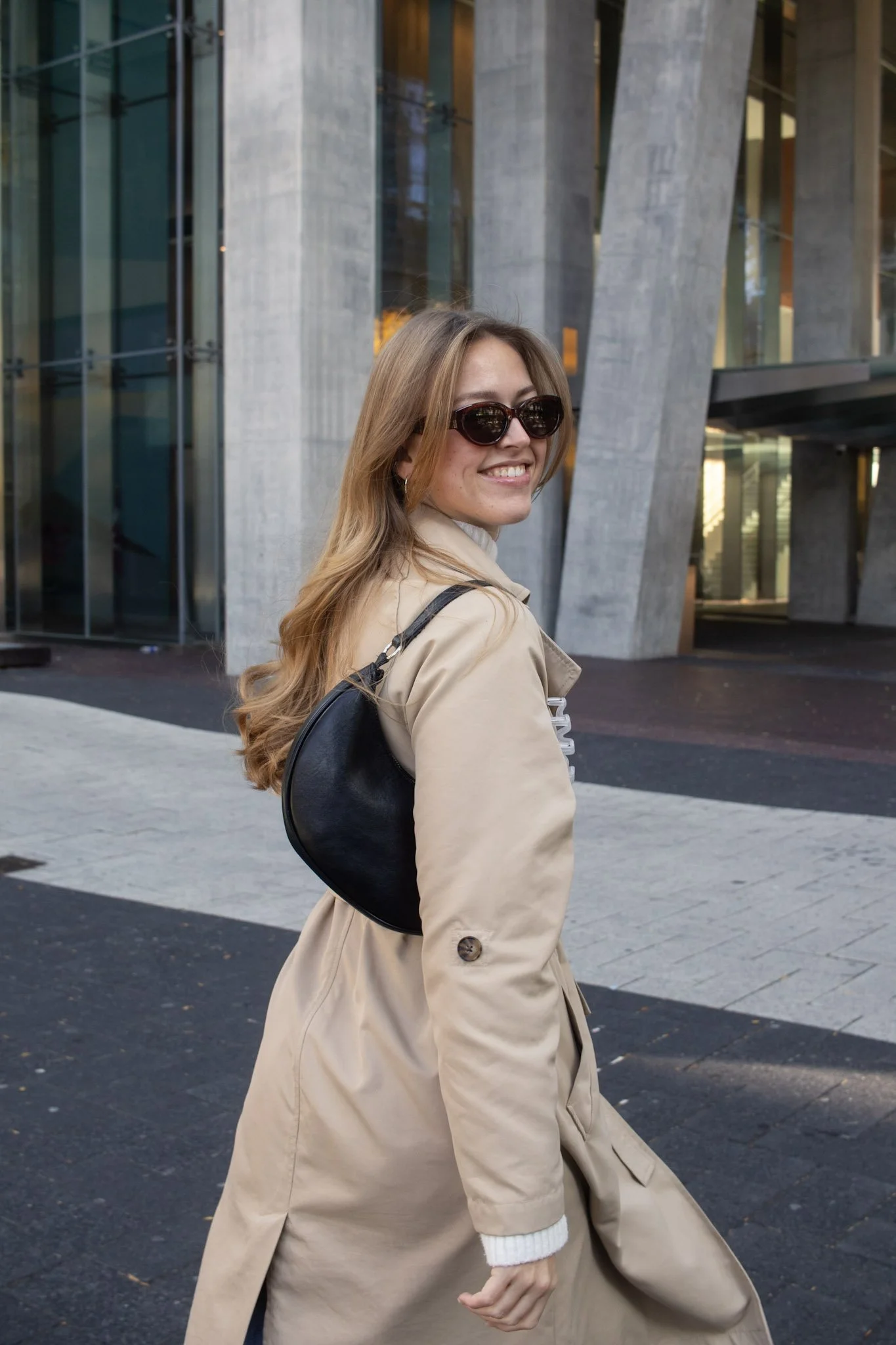 A woman with long blonde hair wearing sunglasses and a beige trench coat, walking outside a modern building with large glass windows and concrete architecture.