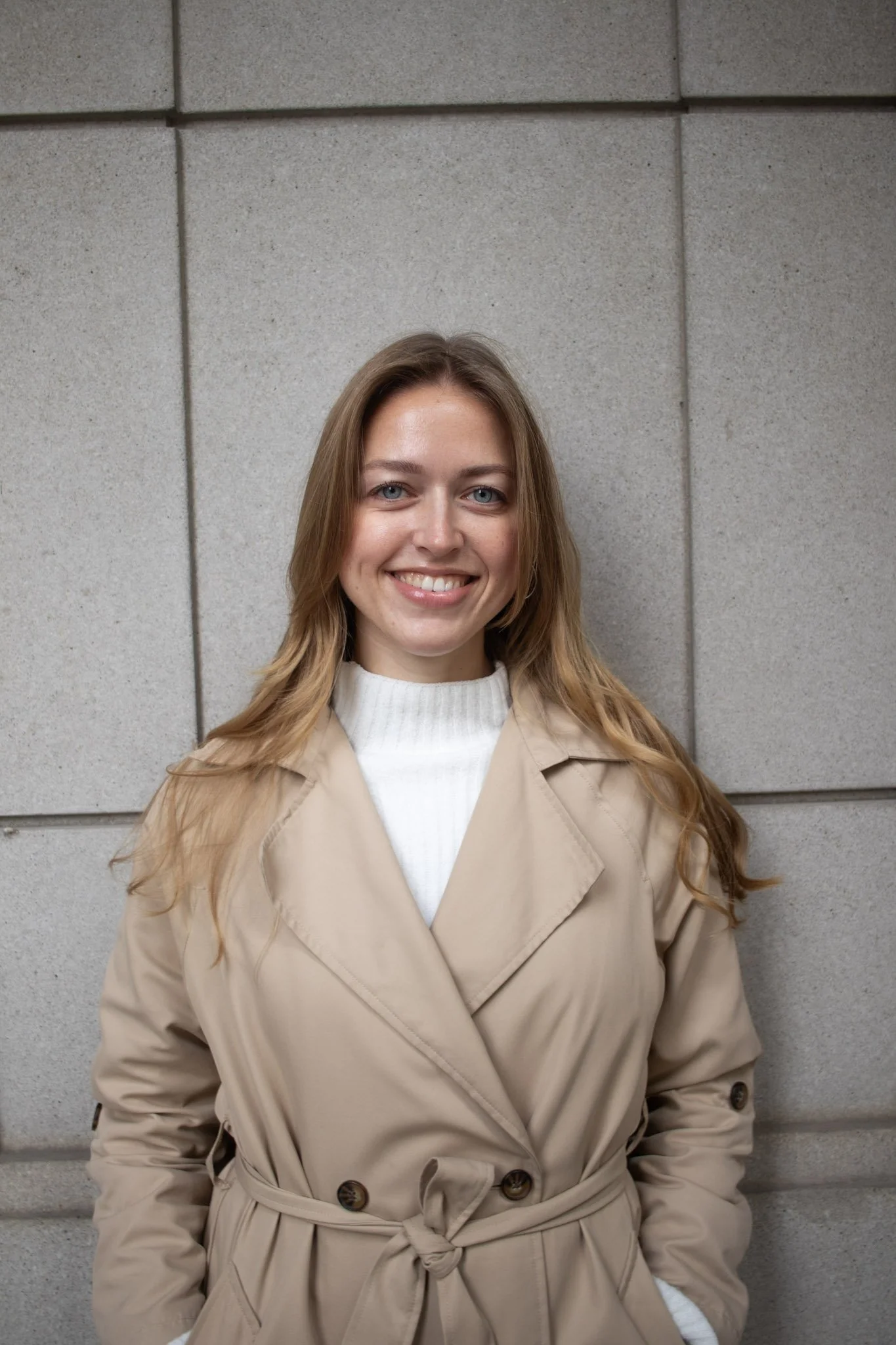 A woman with long blonde hair, blue eyes, and light skin, smiling, standing in front of a gray tiled wall, wearing a beige trench coat over a white turtleneck.