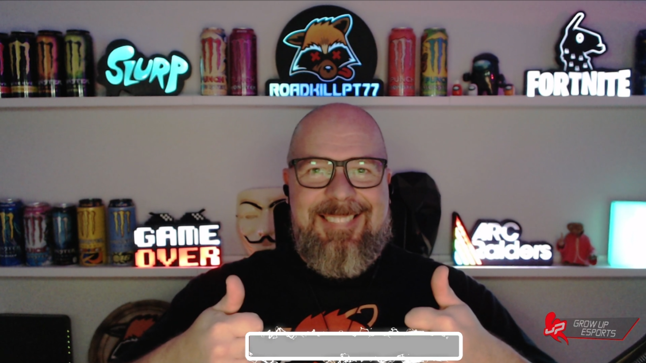 A smiling man with glasses and a beard giving two thumbs up in front of shelves filled with gaming and energy drink cans, neon signs, and gaming merchandise.
