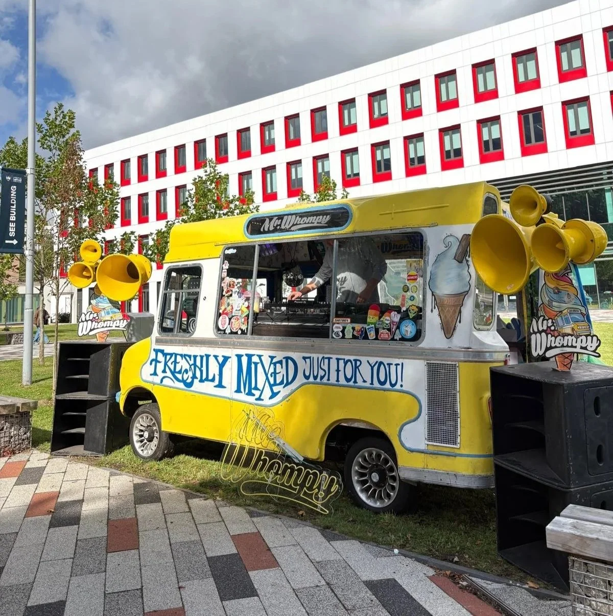 Yellow ice cream truck with speakers, logos, and signs reading "Mr. Whompy" in front of a modern red and white building, with trees and a cloudy sky in the background.