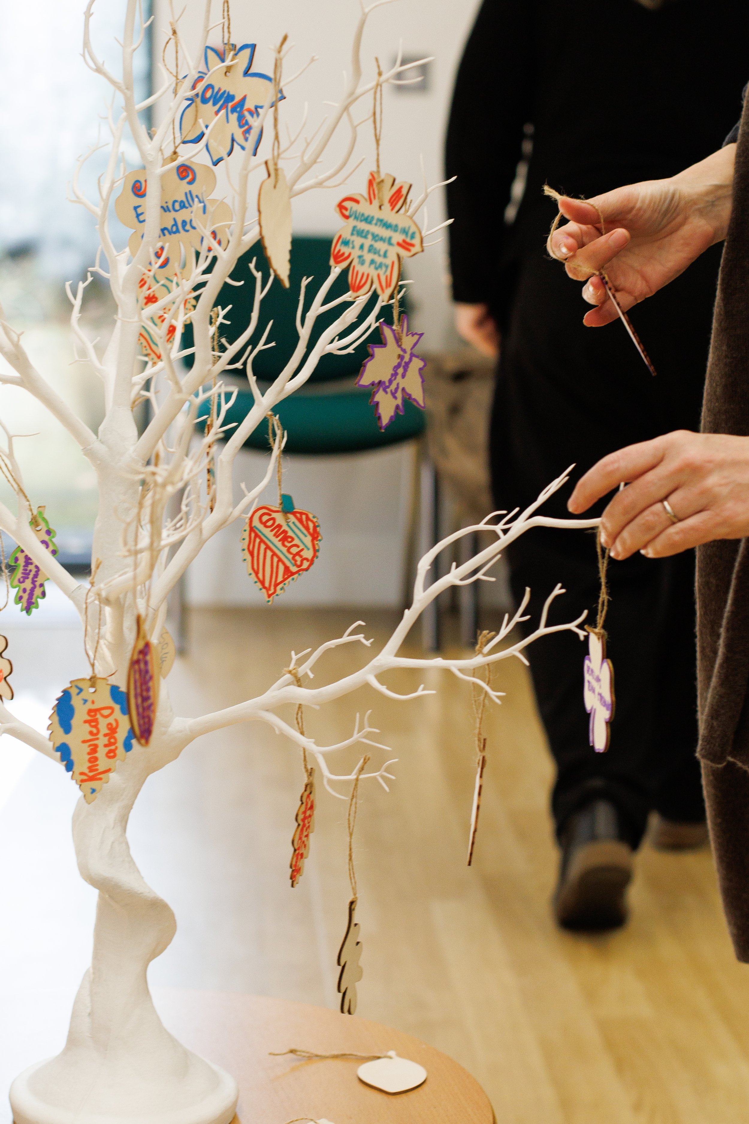 A white decorative tree with colorful paper leaf and oval-shaped ornaments hanging from its branches, situated on a table with a person in the background.