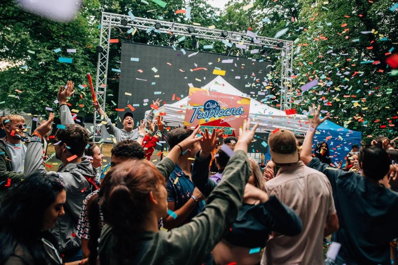 Crowd celebrating at outdoor event with confetti falling, stage with large screen, and tents, in a wooded area.