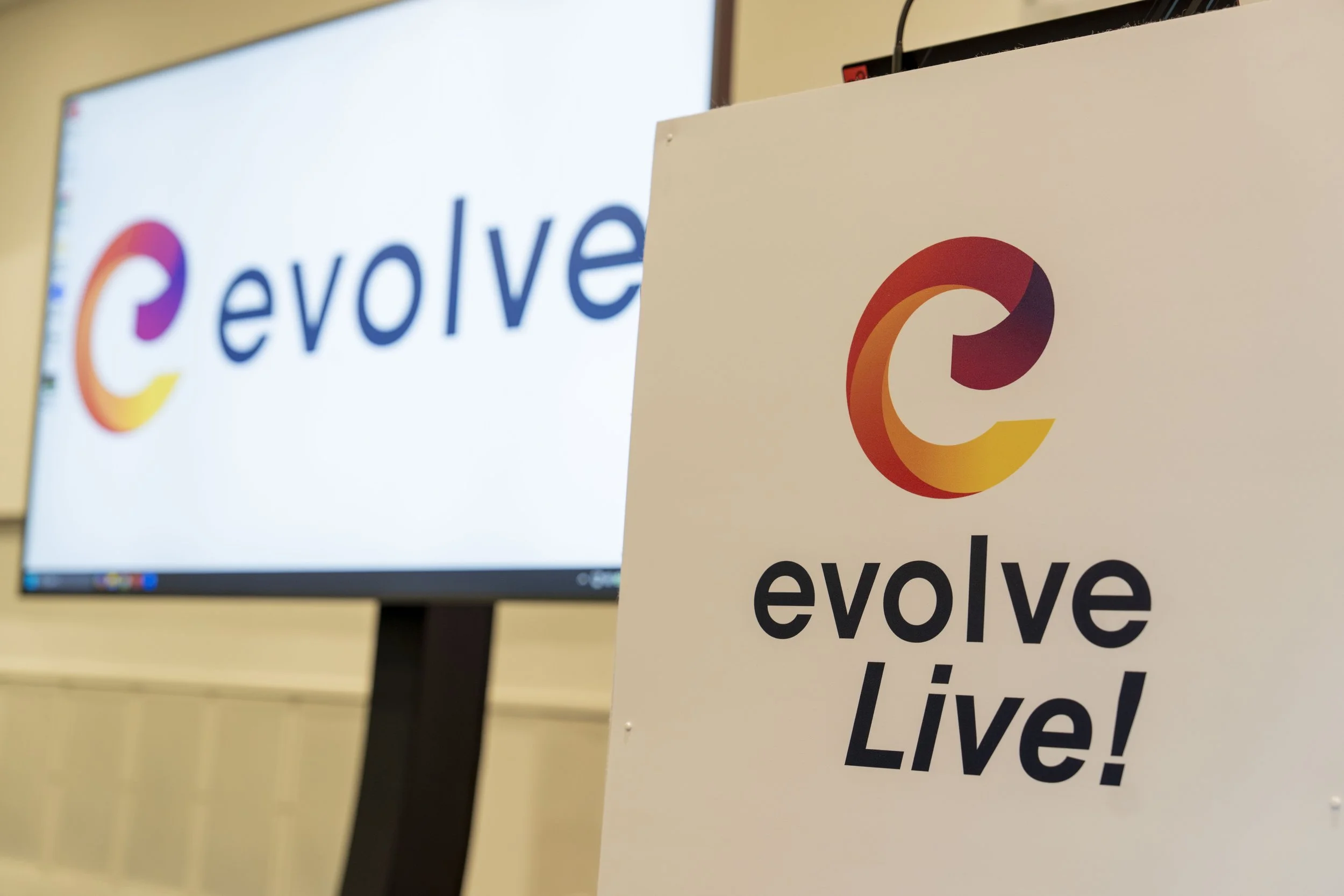 Close-up of a sign with a colorful circular logo and the word "evolve Live!". In the background, a computer monitor displays the same logo