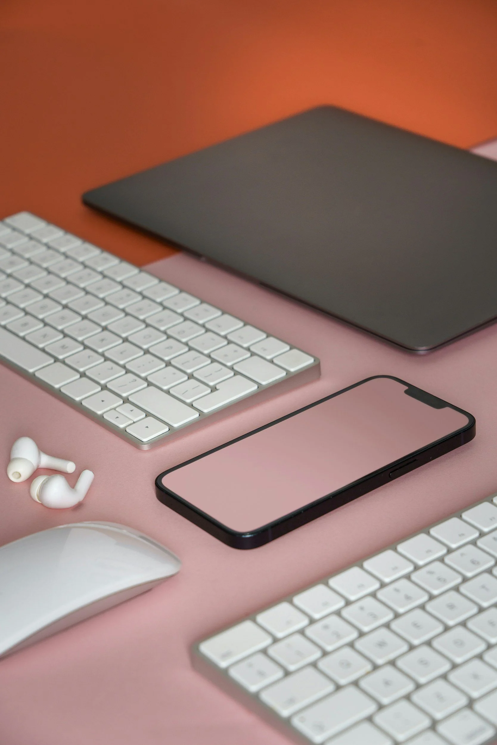 En svart laptop, en vit trådlös mus, en svart smartphone, ett tangentbord, ett par vita trådlösa öronsnäckor på ett rosa bord, bakgrund i orange och rosa.