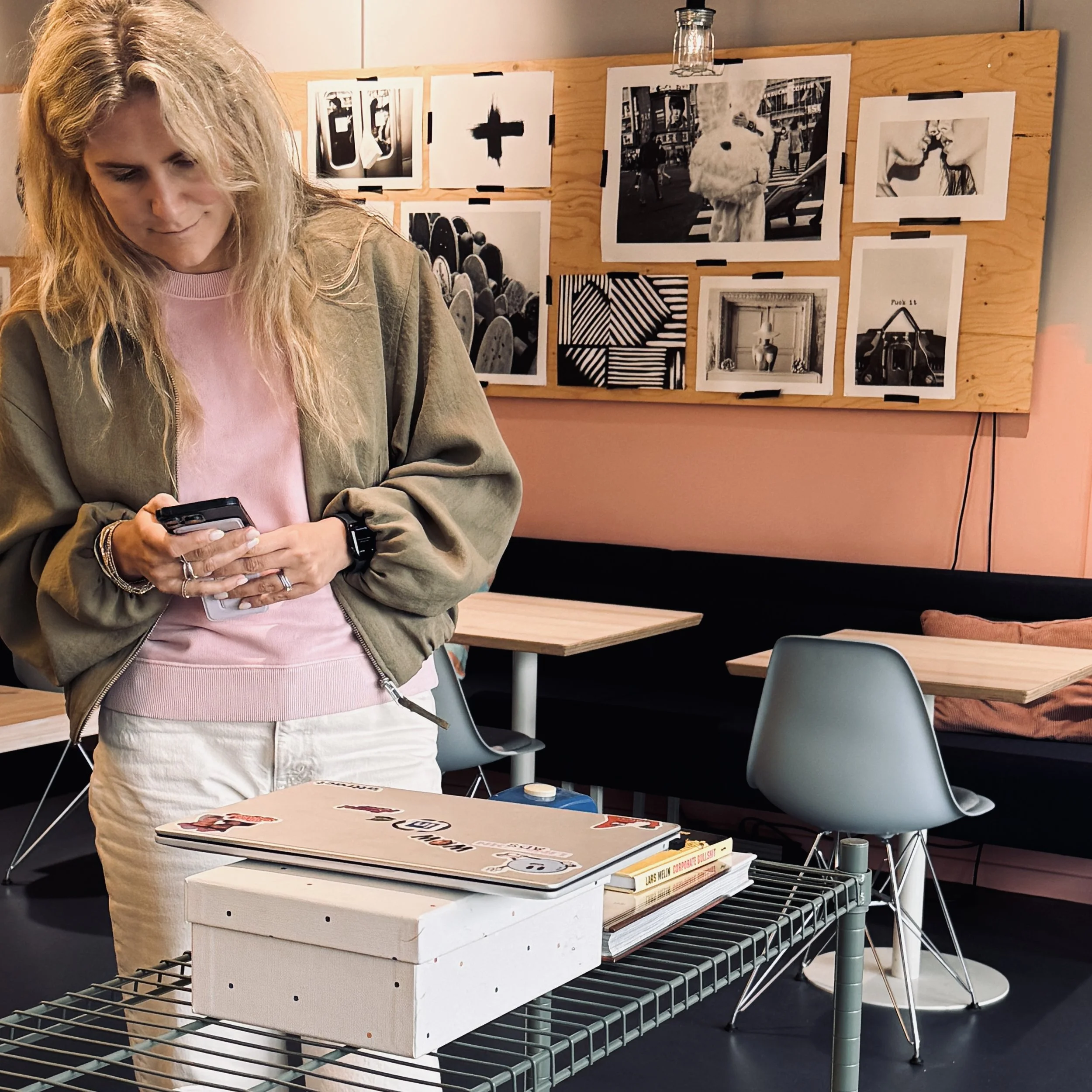 En kvinna med långt blont hår, iklädd en rosa tröja och en grön jacka, står vid en bord med en öppen bok eller katalog. Bakom henne finns en vägg med fotografier och konstverk, inklusive svartvita bilder och en stor bild av en person i ett djur- eller kostymliknande utklädnad.