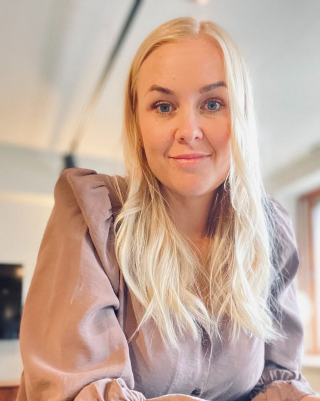 En ung kvinna med långt blont hår och blå ögon, leende, inomhus, klädd i en beige skjorta.