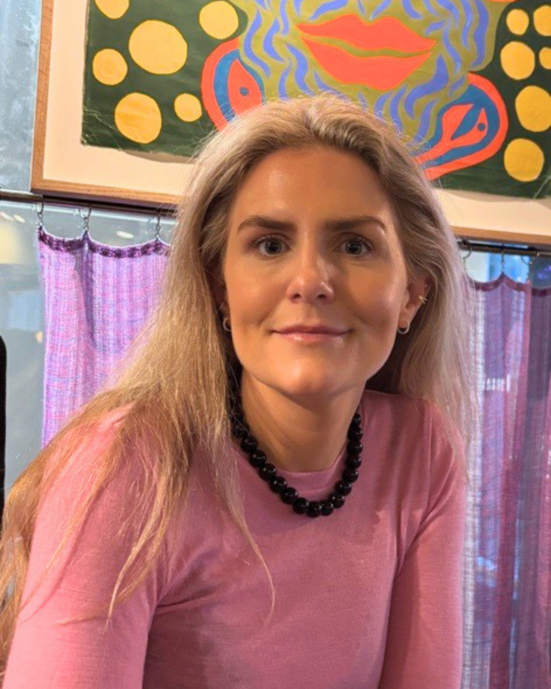 Ung kvinna med långt blont hår, rosa topp, svart pärlhalsband, och små örhängen, står inomhus framför ett färgglatt konstverk och filtar.