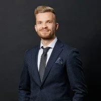 En man i kostym och slips, står mot en mörk bakgrund, ler.