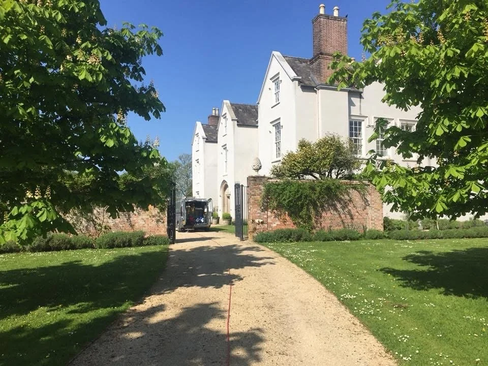 A white house with multiple gables, surrounded by a green lawn and trees, with a gravel pathway leading to the entrance under a clear blue sky.