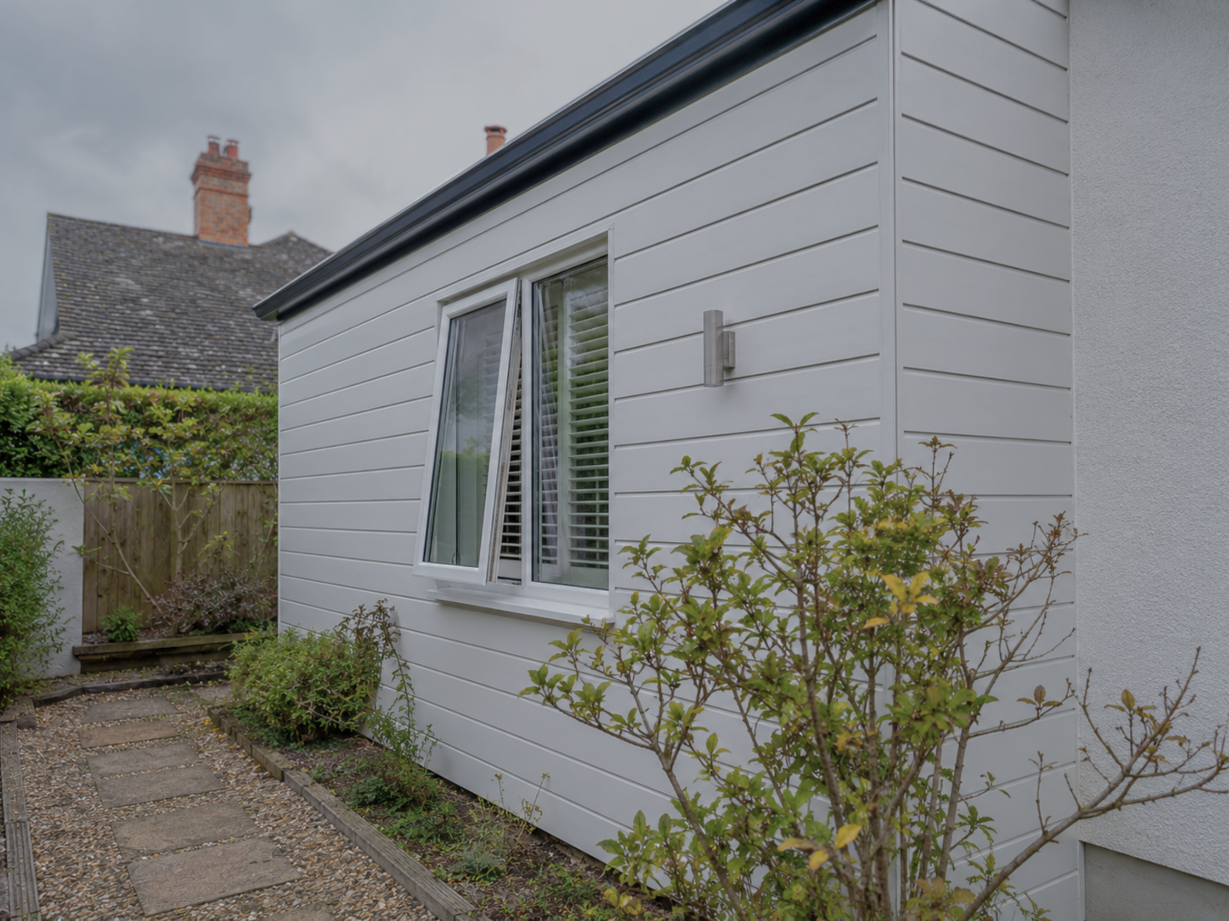 Exterior of a house with white siding, a window. The yard has plants, a small pathway with stepping stones, and a wooden fence. Overcast sky.
