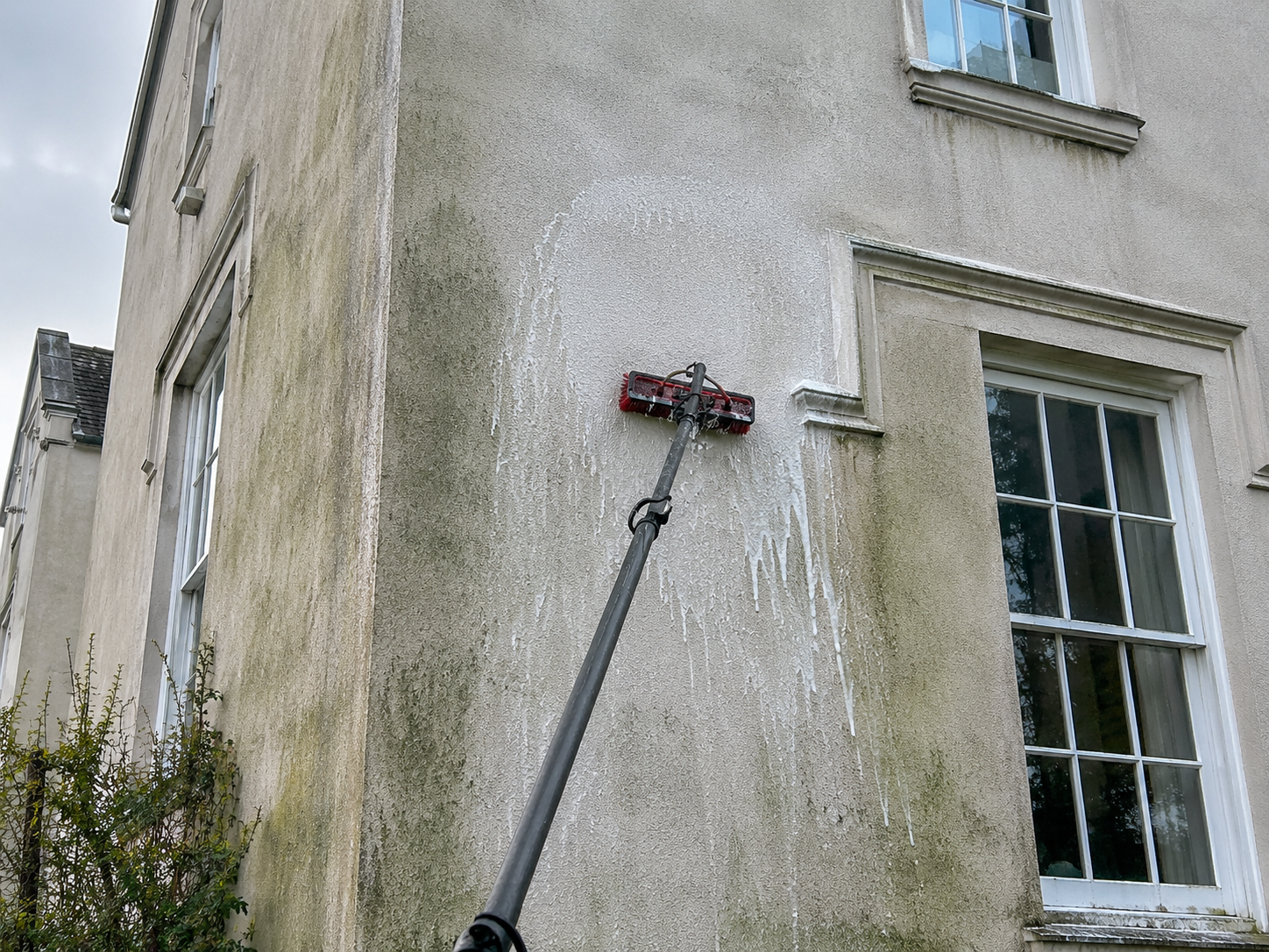 A power washing tool cleaning a dirty exterior wall of a house with window frames.