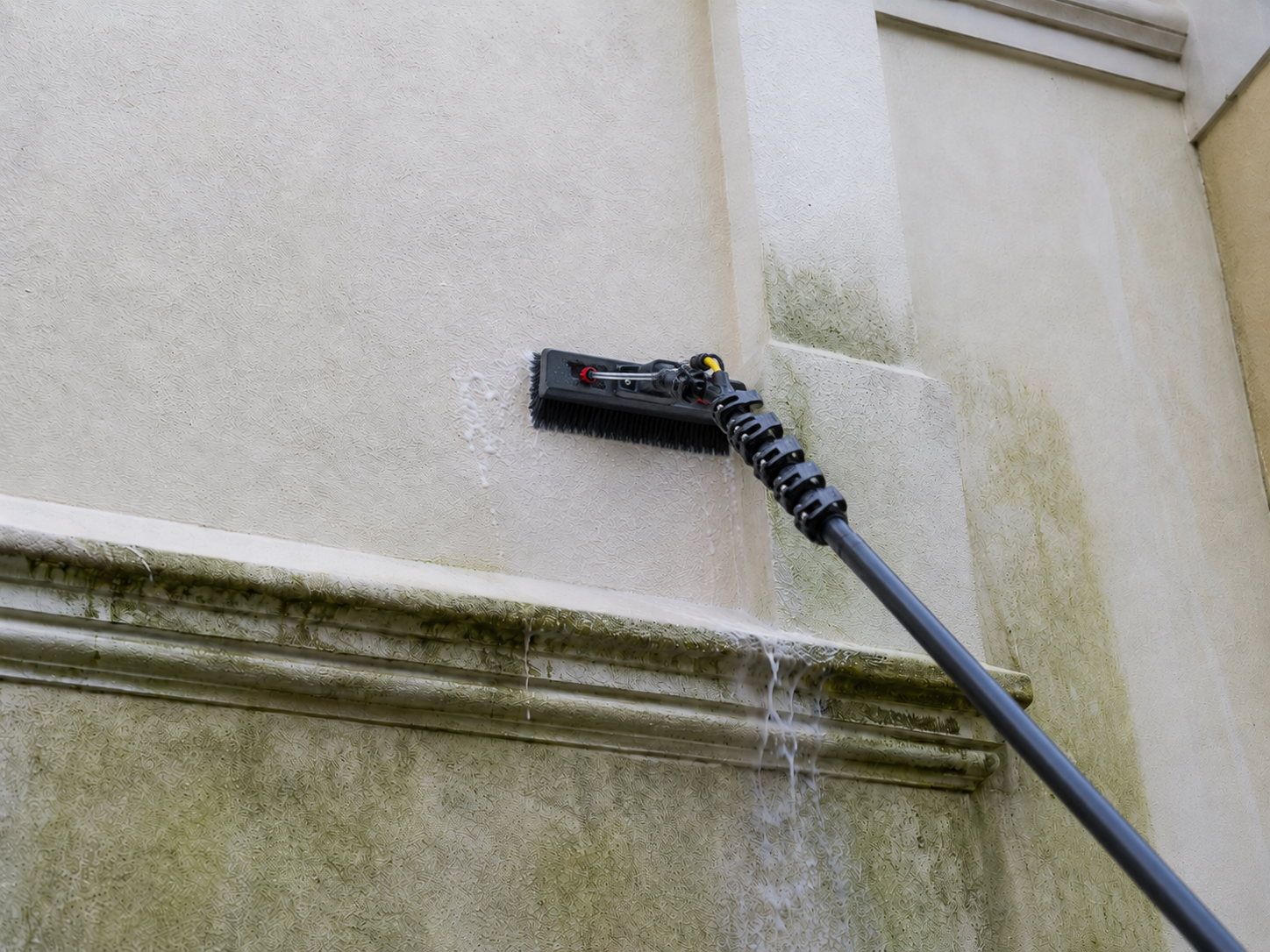 A power washer cleaning a beige exterior wall with soap and water.