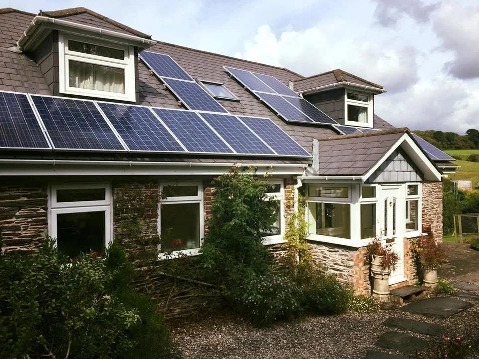 A house with solar panels installed on the roof, surrounded by greenery and cloudy sky.