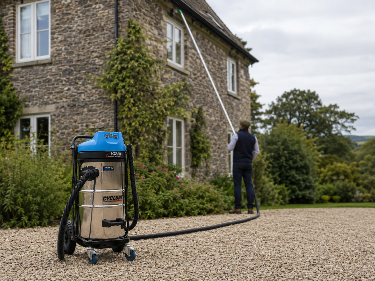 Man cleaning windows of a stone house with a high-reach tool using a cleaning vacuum in front, on a gravel pathway surrounded by greenery.