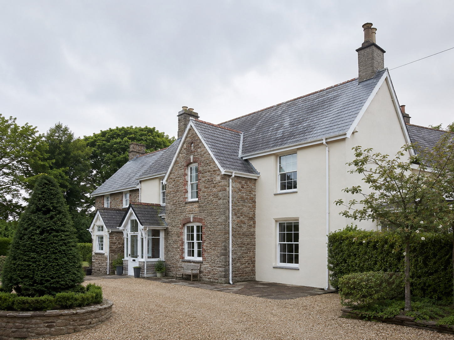 A large house with a combination of stone and off-white siding, multiple chimneys, and white-framed windows, surrounded by greenery and a gravel driveway under an overcast sky.
