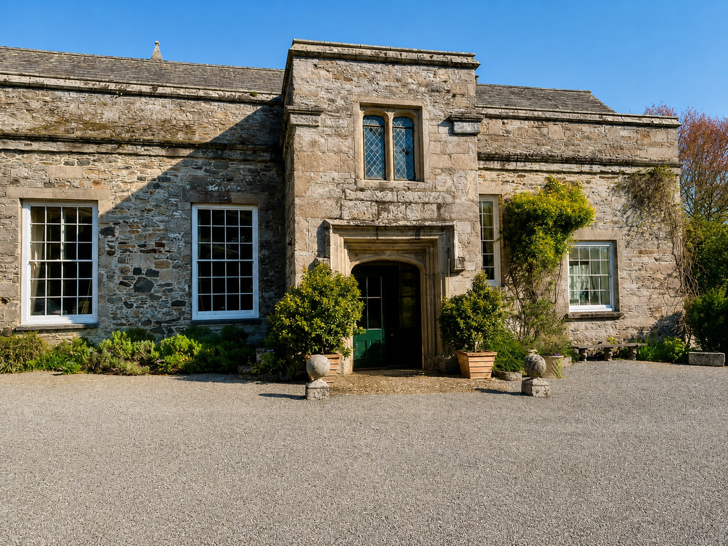 Stone historic building with a dark green door, surrounded by plants and potted bushes, under a clear blue sky.