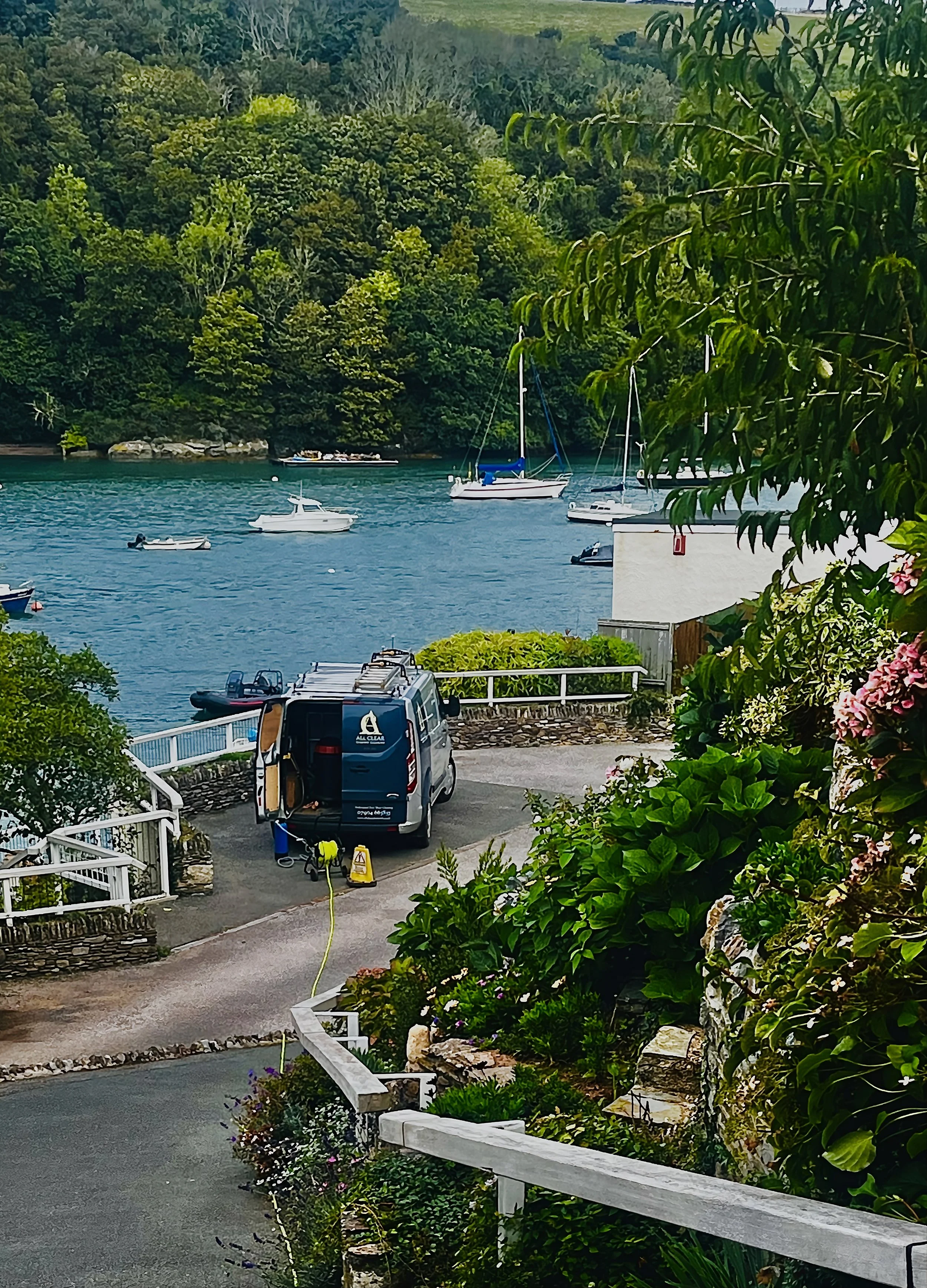A harbor scene with several boats on the water, lush green hills in the background, and plants and flowers in the foreground. A white vehicle is parked near a white railing, with a yellow caution sign and equipment nearby.