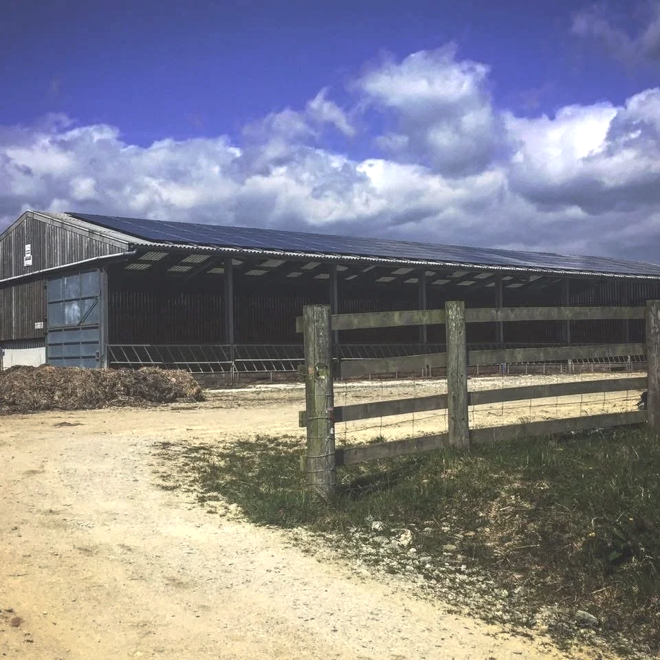 A large metal barn with solar panels on the roof, situated on a dirt farm road with a wooden fence in the foreground, under a partly cloudy sky.