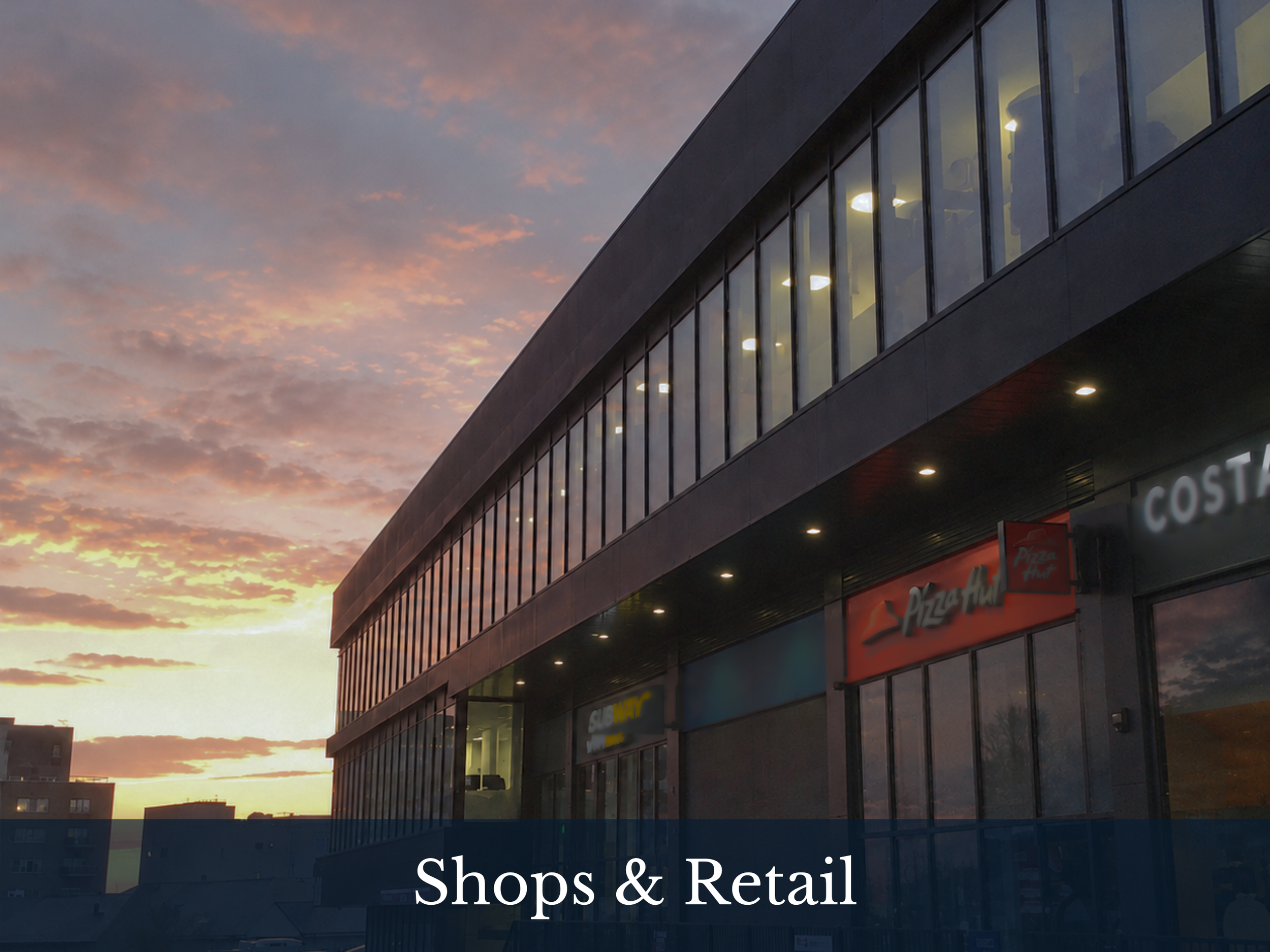 Photo of a shopping mall building with large glass windows, showing store signs like Subway and Pizza Hut, at sunset with a colorful sky.