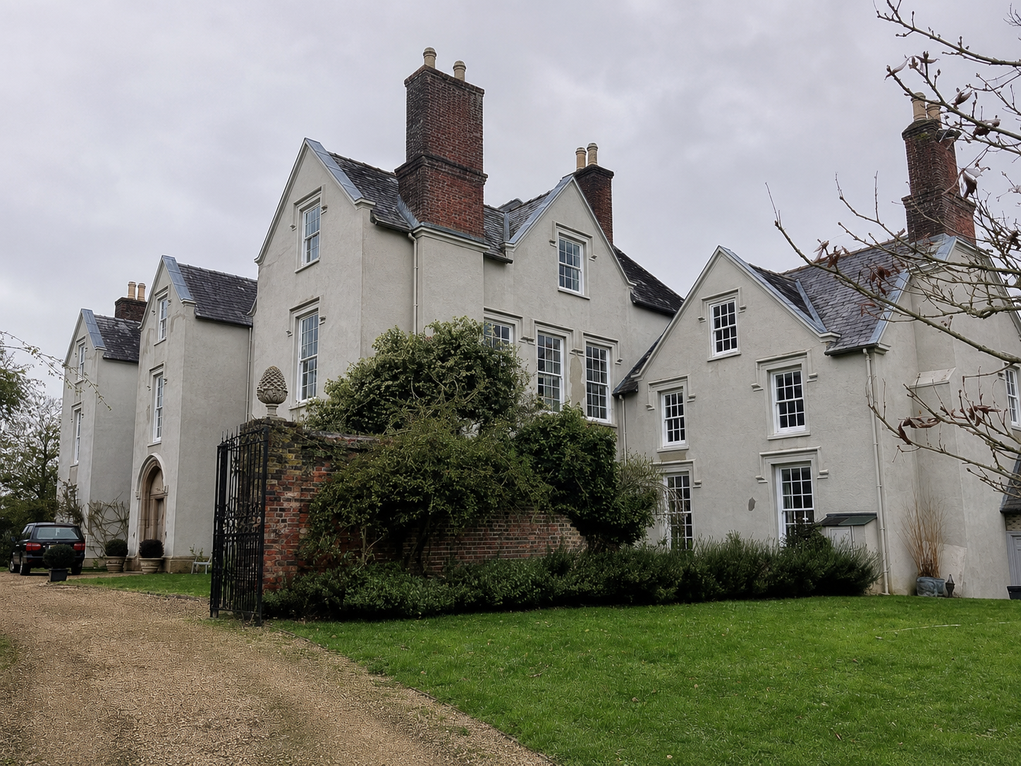A large, historic white mansion with multiple chimneys and tall windows, surrounded by a lawn, bushes, and a brick wall with a black gate, on a cloudy day.