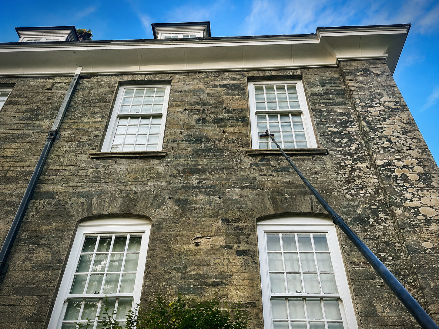 Low-angle view of a brick building with four white-framed windows and a drainpipe on the left side, a long pole cleaning the upper window, and a blue sky with some clouds in the background.