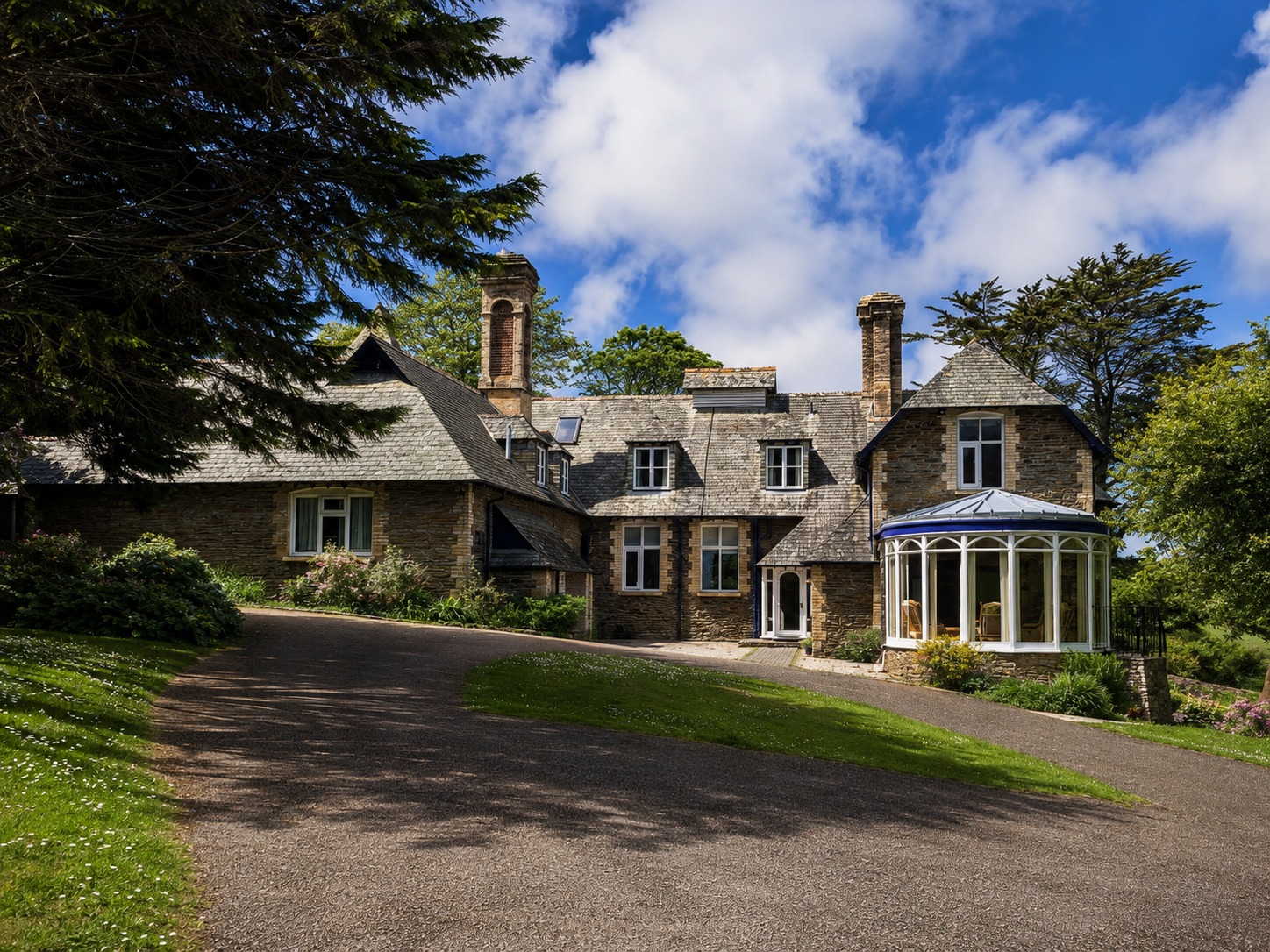A large stone house with a curved glass porch, surrounded by greenery, under a partly cloudy blue sky.