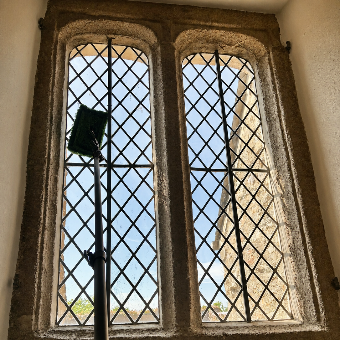 A window with diamond-shaped iron bars, an upright window cleaning tool with a green rectangular cleaning pad attached, visible from inside a stone and plaster building.