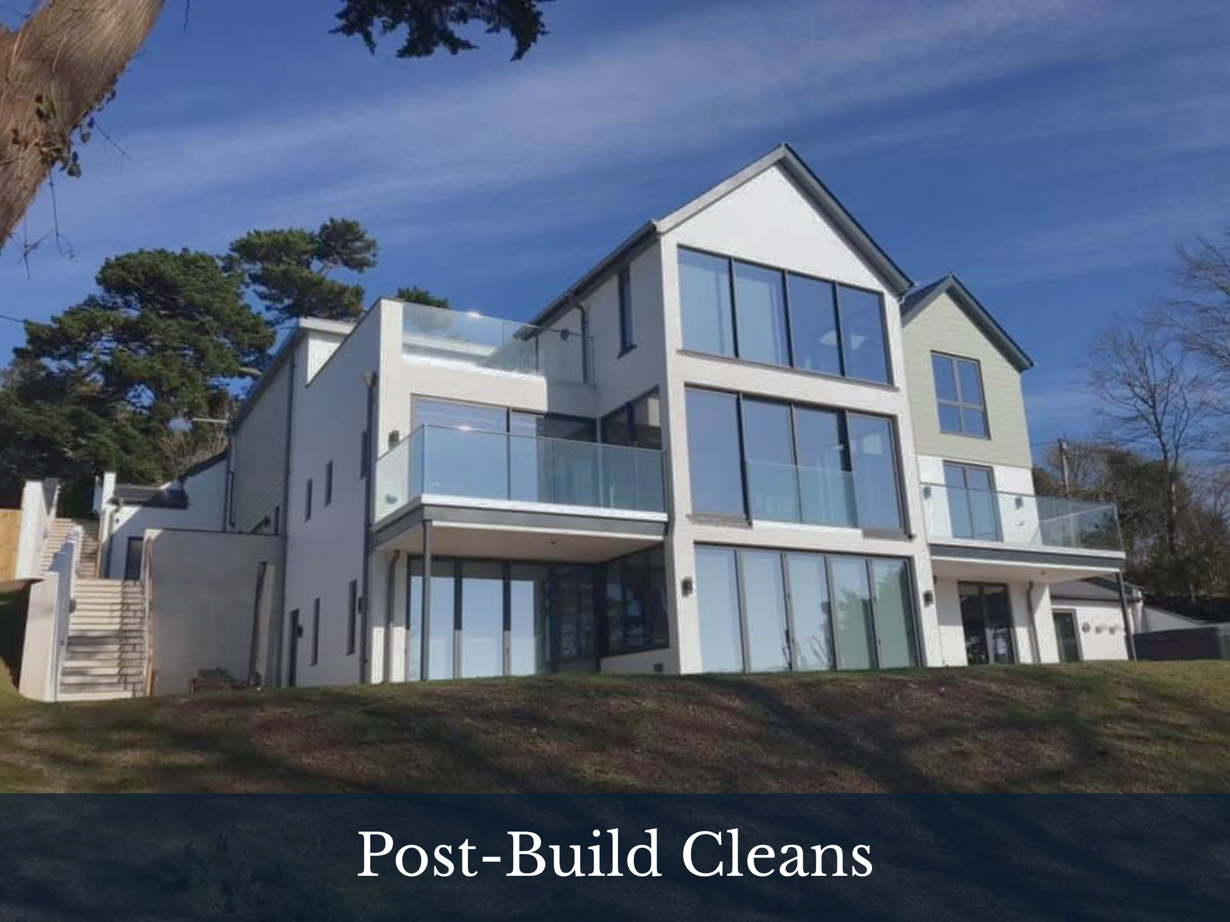 Modern white multi-story building with large glass windows and balconies, surrounded by trees and a clear blue sky.